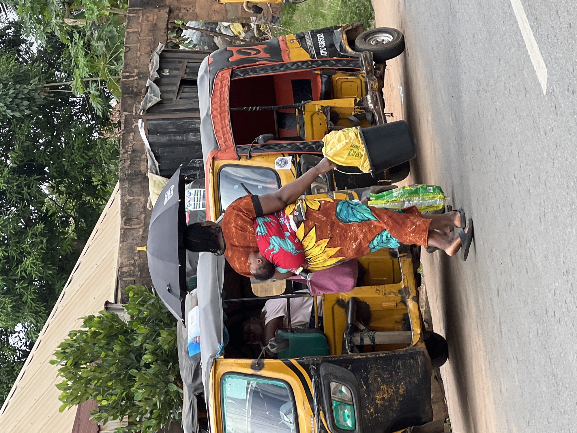 This image captures a vibrant daytime street scene in Obosi, Nigeria, featuring a woman with a baby, a motorized tricycle, and various elements of the local environment.
The central subject is a woman standing next to a yellow and black motorized tricycle, commonly known as a "keke napep." She is wearing a colorful, patterned dress, likely an African print, with prominent red, orange, and blue hues and a large yellow sunflower motif. A baby is securely strapped to her back, nestled against her in the same patterned fabric. The woman holds a black umbrella in her left hand and a black bucket, containing a yellow bag, in her right. She is wearing dark sandals. At her feet, or resting on the tricycle, are several green and white bags, one of which visibly says "MAMA'S PRIDE."
The tricycle has a yellow body with black accents and a red interior. Text "ATRIC A36500" is visible on its side. Another individual, possibly the driver or a passenger, can be seen partially inside the front of the tricycle, wearing a white shirt. A small white sticker on the front of the vehicle displays the word "KIND."
The background shows a paved road with a white lane marker. On the left, lush green trees with dense foliage are prominent, along with parts of a building featuring a corrugated metal roof and brickwork. The scene depicts an ordinary moment of daily life and transportation in the area.