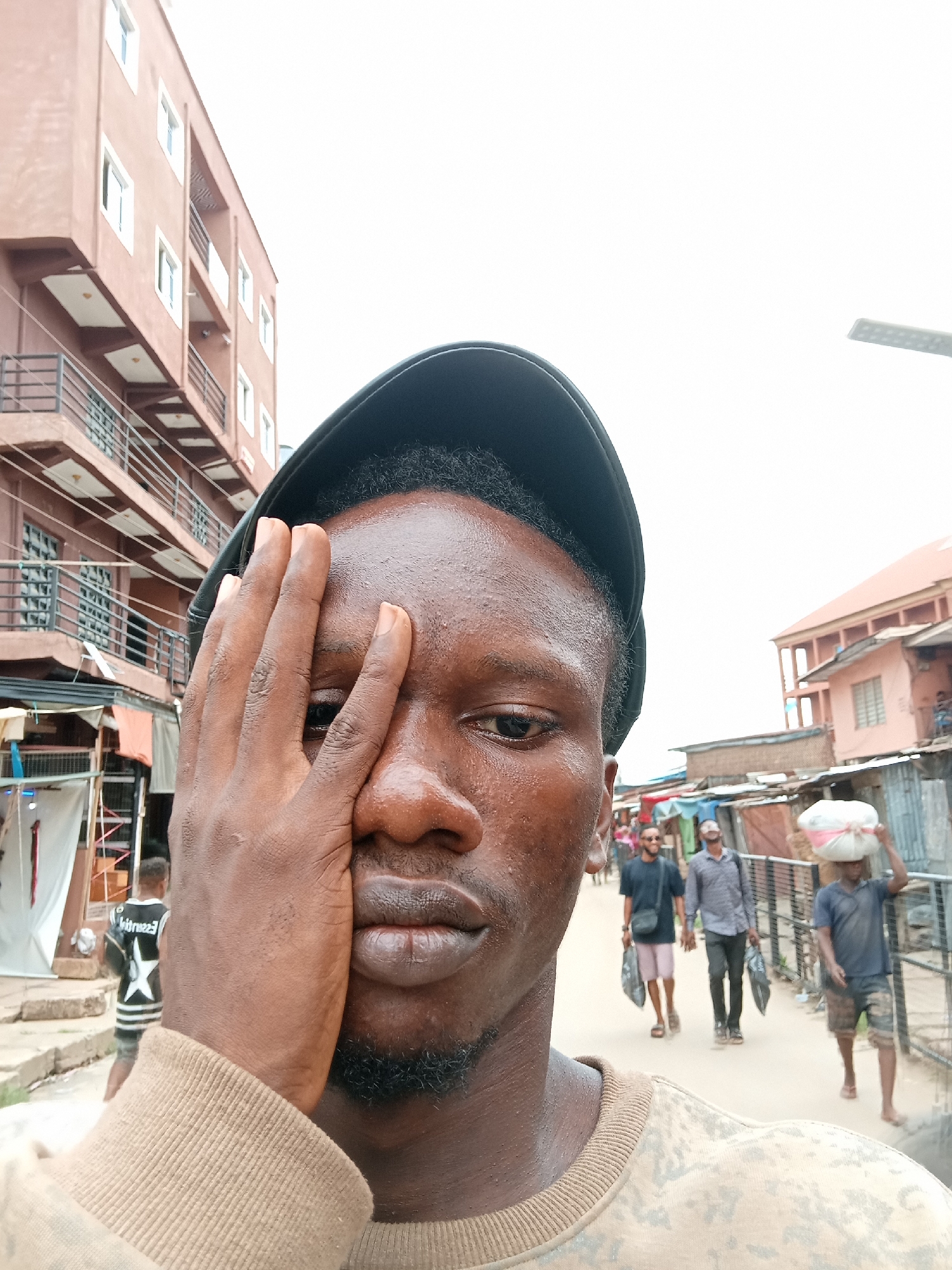 This image captures a close-up selfie of a young man against the backdrop of a busy street scene in Onitsha, Nigeria, during daylight hours. The main subject, with dark skin, occupies the foreground, wearing a black baseball cap and a light brown or camouflage-patterned long-sleeved top. He has raised his right hand to partially cover his face, obscuring his nose and mouth, leaving his left eye visible. His skin appears to have a sheen, suggesting warmth or humidity. A short, dark beard is visible on his chin.
In the midground and background, the street is populated with several other men walking. One man in the distance is seen carrying a large white bag on his head, while others carry black plastic bags in their hands. The urban environment features a multi-story building with reddish-brown walls and balconies on the left side of the frame, contrasting with more informal structures, possibly market stalls or smaller residential buildings constructed with corrugated metal and painted walls, on the right. The sky overhead is bright, suggesting a clear day or overexposure.
A notable detail is visible text on a black and white striped jersey worn by a person walking in the background on the left; the word "Wessentials" or a similar term can be partially discerned. The overall scene depicts a slice of daily life on a vibrant street in a Nigerian city.