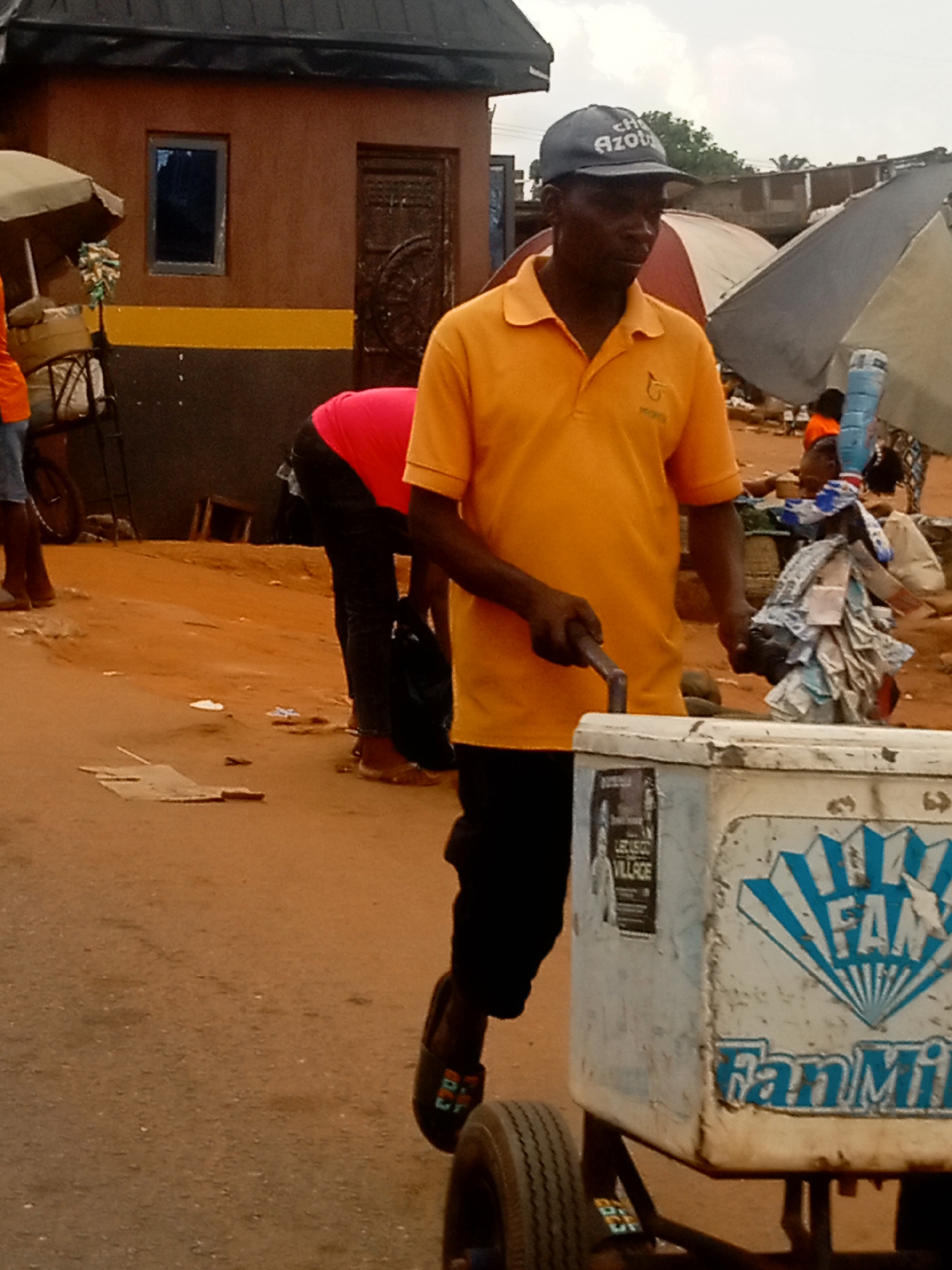 In Ogidi, Nigeria, a man wearing an orange polo shirt and a dark cap pulls a wheeled cooler with "Fan Milk" printed on its side. He is on a dirt road amidst scattered debris. A woman in a pink shirt is bending over nearby. There are other people and vendor umbrellas in the background, and a building with a dark-colored facade is partially visible. The sky is overcast.