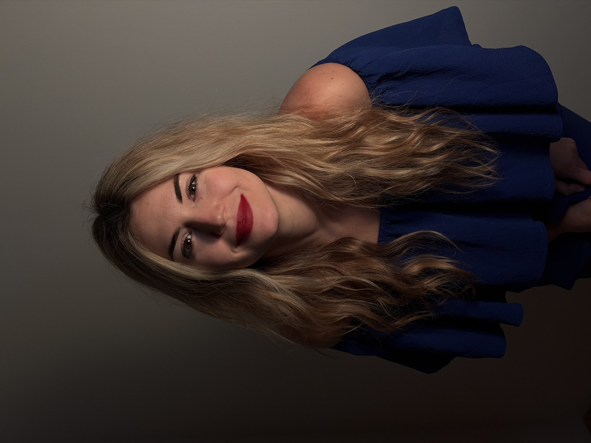 A single adult female is depicted in a head-and-shoulders portrait, positioned against a plain, solid-colored wall that transitions from light grey on the right to dark grey on the left due to localized lighting. The subject has long, wavy blonde hair with visible darker roots, cascading over her shoulders. She wears a dark blue, off-the-shoulder garment featuring ruffled fabric along the neckline. Her face is directed towards the camera, illuminated primarily from the viewer's upper right, creating a shadow gradient across her face and the background. Her eyes are dark brown and gaze directly forward. Her lips are coated with red lipstick, and she exhibits a slight smile. A small, dark mark is visible on her left cheek, below the eye. Her shoulders are bare, and her hands are partially visible at the bottom of the frame, in a clasped or resting position. The scene is located in the city of Budva, Montenegro.