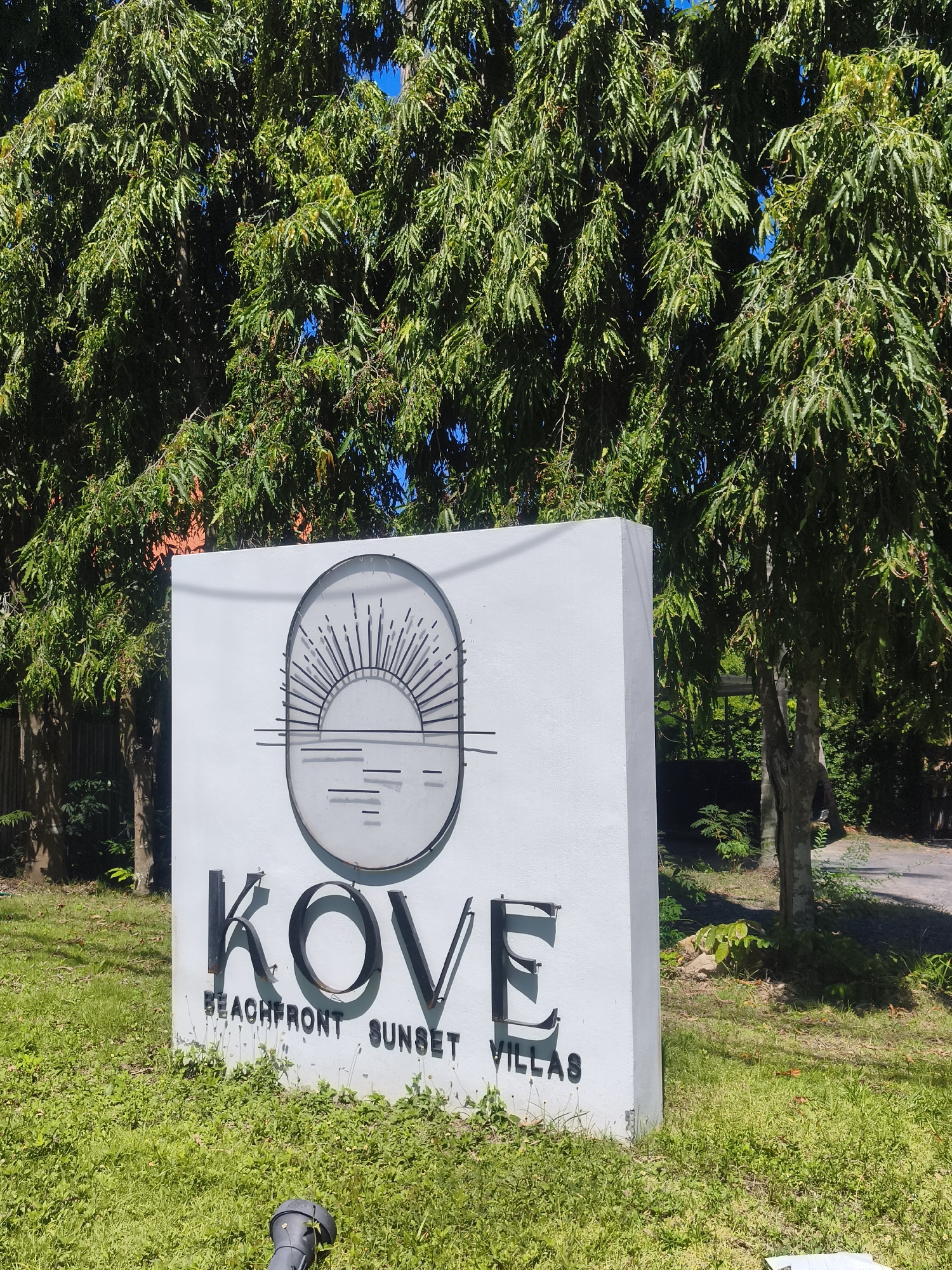 In the city of Baan Thong Po, Thailand, a white rectangular sign stands on a grassy area, partially obscured by lush green foliage from overhead trees. The sign displays the name "KOVE" in large, black, metallic letters. Below "KOVE," in smaller black text, are the words "BEACHFRONT SUNSET VILLAS." To the left of the text, the sign features a circular emblem containing a stylized depiction of a setting sun over water, rendered with thin, dark lines. The environment is sunny and appears to be an outdoor setting, likely a public space given the sign.