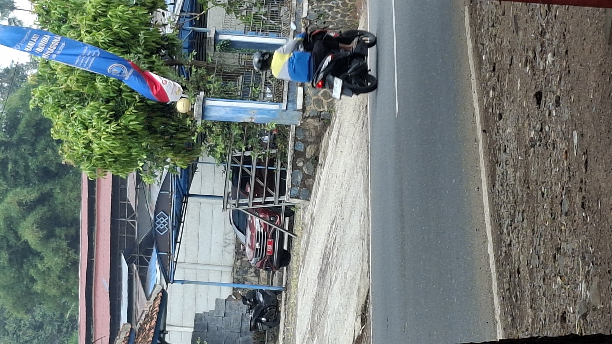 The image captures a scene on a roadside in Cilaku, Indonesia. In the foreground, a paved road with a white line marking its edge is visible. The middle ground features a slope leading up to a property. A person is riding a dark-colored motorcycle on the road, wearing a helmet and a blue and yellow jacket. To the left, a red SUV is parked, with a black motorcycle beside it. The background shows a building with a red roof, a blue awning, and lush green trees. A blue and white flag is also prominently displayed. The overall environment appears to be an outdoor, semi-urban setting.