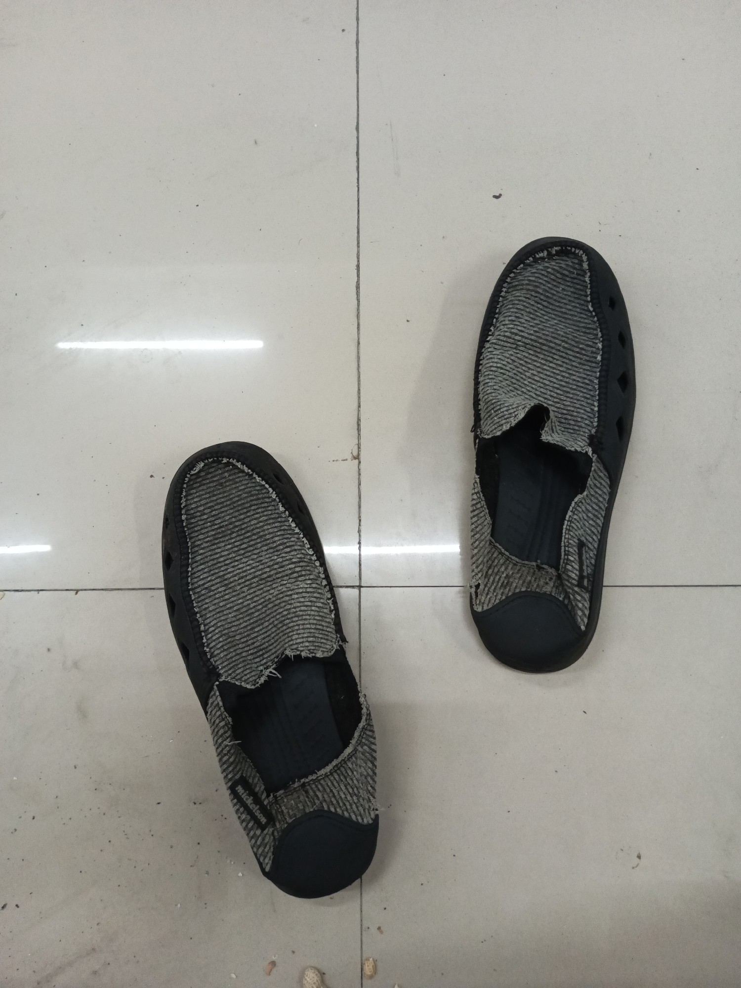 Two black and grey slip-on shoes with holes in the fabric upper are placed on a light grey tiled floor. The shoes are oriented in opposite directions, with the left shoe pointing towards the bottom left and the right shoe pointing towards the top right. The tiles are arranged in a grid pattern, and there are faint reflections from overhead lights on the floor. The overall scene appears to be indoors, possibly in a home or commercial space. There is no indication of the city of Makassar, Indonesia, or any other specific location.