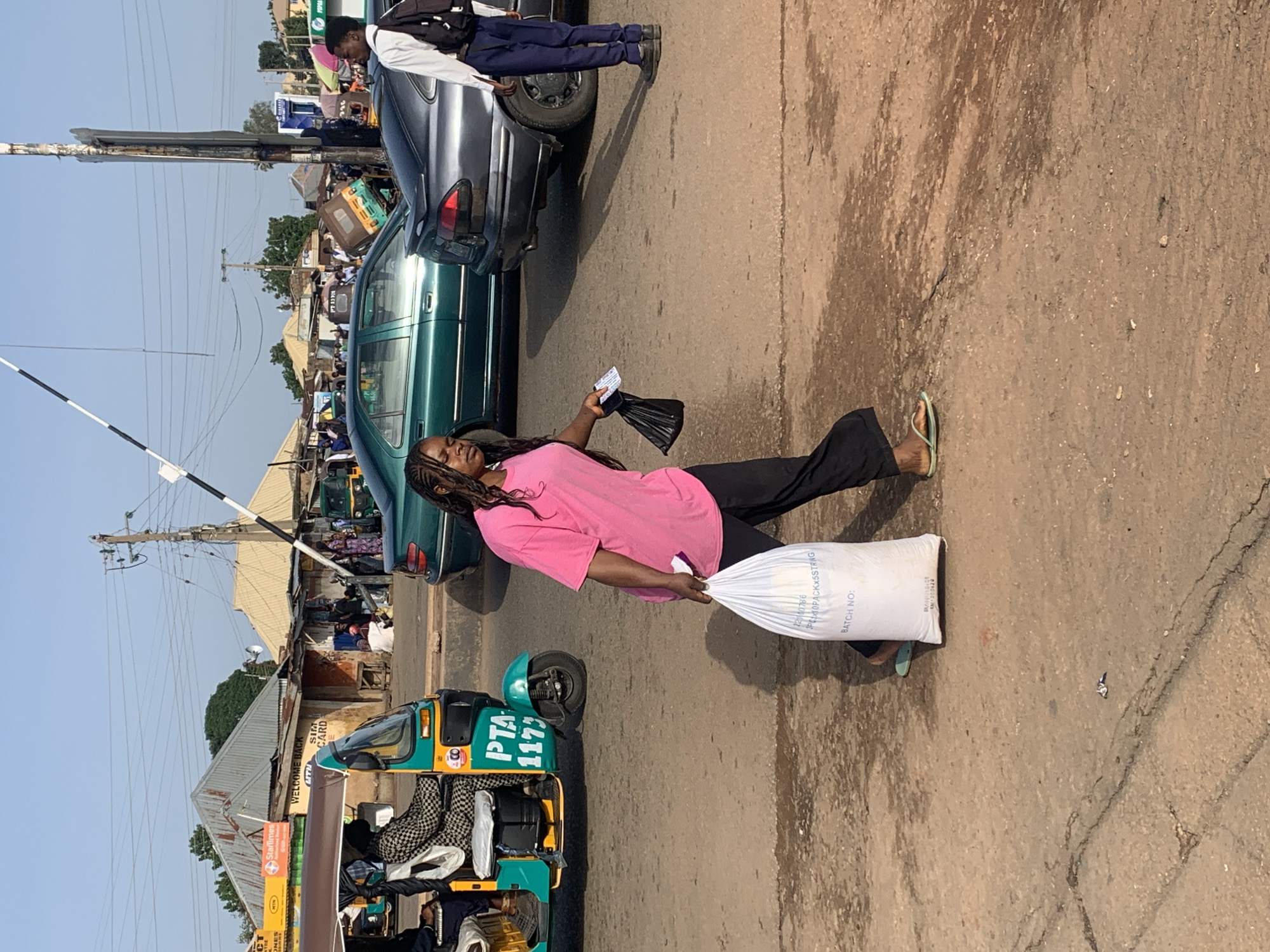 In the city of Gura Top, Nigeria, a young woman with braided hair, wearing a bright pink t-shirt and black pants, walks across a dusty road. She carries a large white bag filled with what appears to be goods, along with a black plastic bag in her other hand. Behind her, a green car and a grey car are stopped, suggesting traffic or a waiting area. A motorized tricycle, a common mode of transport in Nigeria, is visible to her left. The background shows a bustling street scene with small shops, market stalls, and utility poles, indicative of a lively urban environment. The sun casts long shadows, indicating it is likely late afternoon.