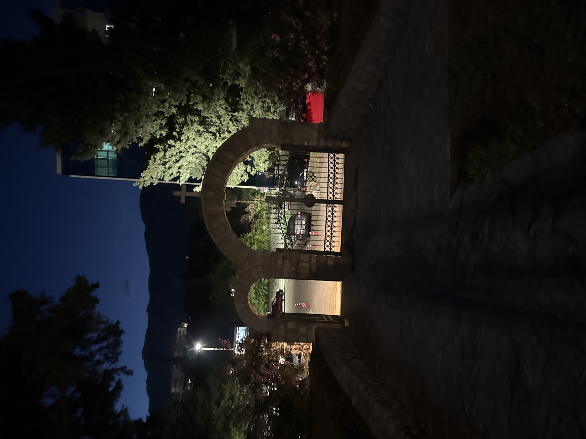 The image captures an entranceway at dusk in Boreti, Montenegro. A cobblestone path leads towards a stone archway with a wrought-iron gate. Beyond the gate, a red car is visible, along with other vehicles and parked cars in the distance. The scene is illuminated by streetlights and the faint glow of the night sky. Trees flank the entrance, and in the background, a modern building stands tall against the silhouette of mountains. The overall ambiance is serene, with a hint of urban life integrated into the natural landscape.