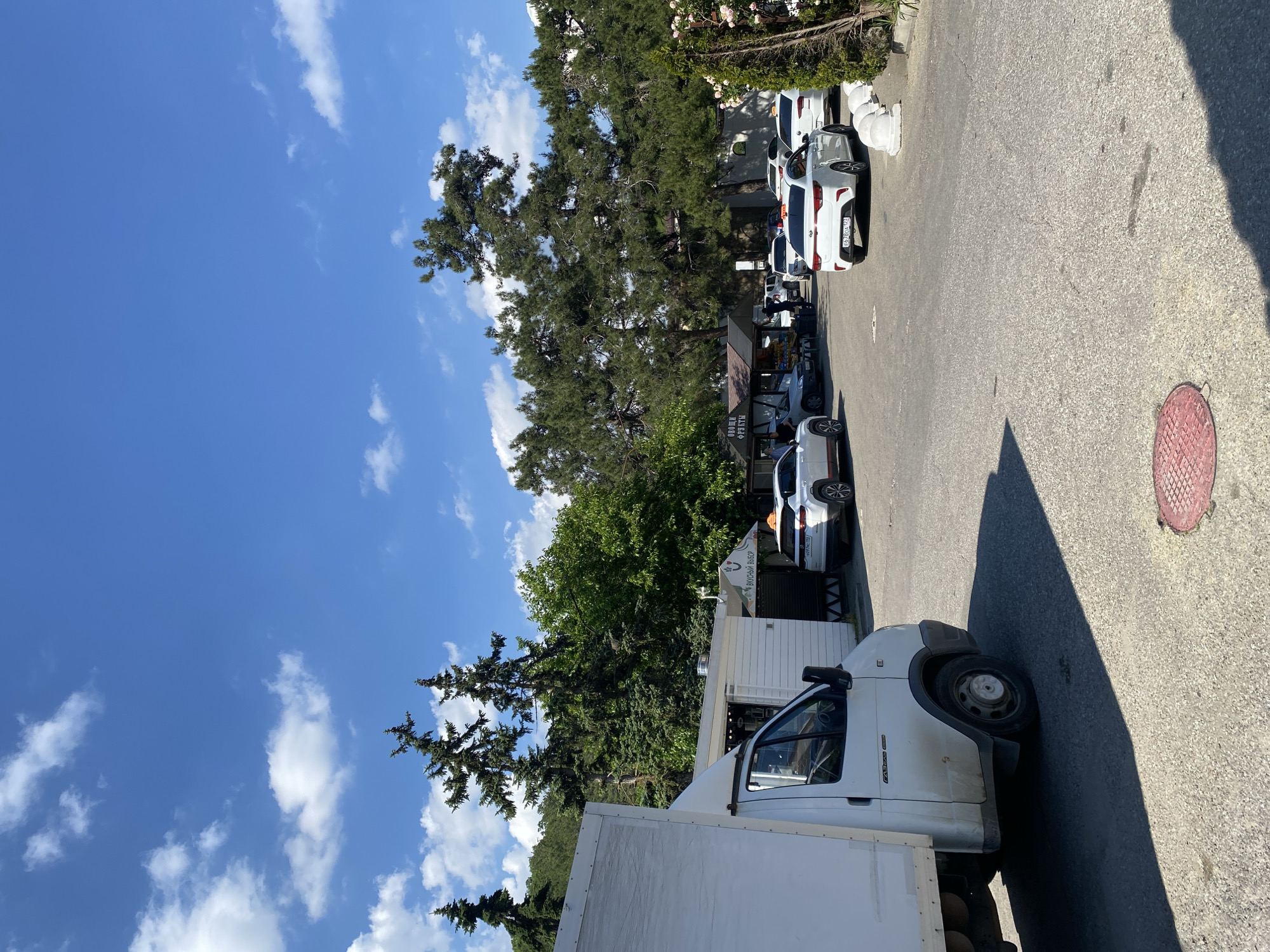 A daytime street scene unfolds under a bright blue sky dotted with fluffy white clouds. The image is taken from a slightly elevated perspective, looking down a paved road or driveway lined with trees and parked vehicles.
In the foreground, the front cabin and cargo box of a white delivery van are prominently visible on the left, facing right. Its front wheel and a deep shadow cast on the asphalt are discernible. The van appears to be a Fiat Ducato or a similar model, with faint branding visible on its side.
Along the right side of the road, several cars are parked bumper-to-bumper. A white sedan, possibly a Kia K5 or similar, is clearly visible, with its Russian license plate displaying "082 A 001" and "23" (likely a region code for Krasnodar Krai, consistent with Abrau-Dyurso). Further down the road, another white car and parts of others are parked.
Behind the parked cars and bordering the road are tall, dense coniferous trees, possibly pines or cypresses, casting long shadows. Interspersed among the trees are glimpses of buildings or small commercial stalls. One stall features a sign with Cyrillic text, partially reading "ОВОЩИ ФРУКТЫ" (Vegetables Fruits), suggesting a fresh produce stand. Another sign above a white awning reads "ВКУСНЫЙ ВЫБОР" (Delicious Choice), indicating another food or beverage establishment.
The ground is asphalt, with a red circular manhole cover visible on the right side of the road further down. The strong sunlight and clear shadows suggest it is midday or early afternoon. The overall atmosphere is calm and typical of a sunny day in a small town or resort area.