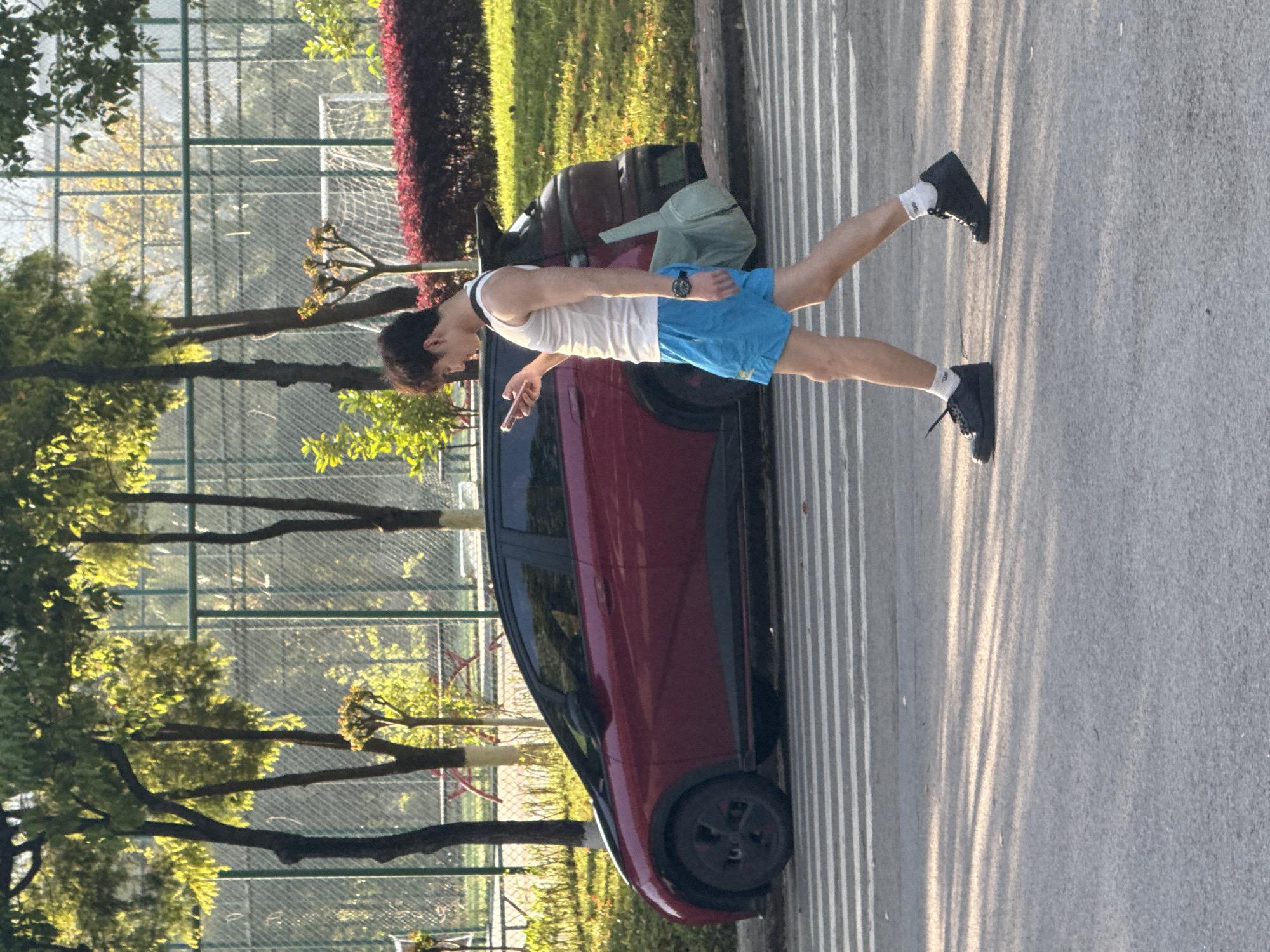A horizontal, eye-level shot shows a person standing next to a red car. The person is wearing a white tank top, blue shorts, white socks, and black shoes. The person appears to be holding a cell phone and looking at the phone. The red car is parked on an asphalt road next to a curb. Behind the car, there are trees, grass, and a fence. In the background, the landscape suggests the location is Jiangjin, China. There is a bag on the roof of the car. The day is sunny.