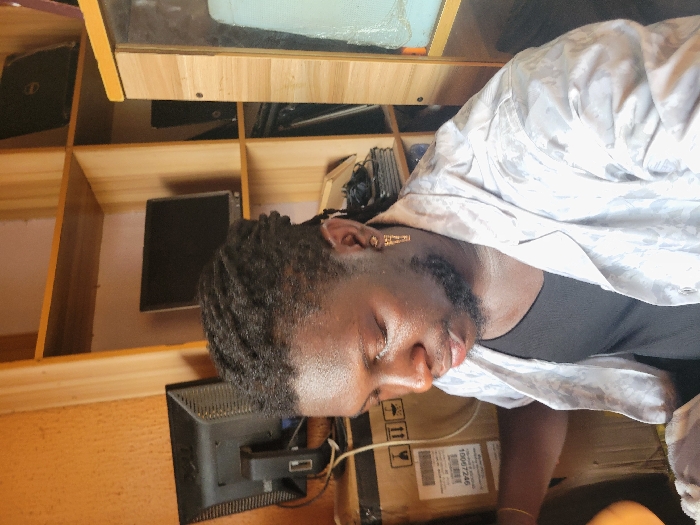 The image displays a male individual in an indoor setting within Makurdi, Nigeria, focusing downwards. The subject has dark skin, short dreadlocks, and a trimmed beard. He wears a black undershirt beneath an unbuttoned, light-colored collared shirt with a subtle pattern. A gold-colored earring, possibly an ankh or similar symbol, is present in his right ear, and a gold-toned bracelet adorns his left wrist.
The background consists of a light-brown wooden shelving unit. The top shelf holds two black laptops, one partially visible. The middle shelf displays two black desktop monitors. The bottom shelf contains stacked black electronic components. To the subject's left, a large cardboard shipping box is visible, featuring labels including "10007246" and handling symbols, with a black cable running alongside it. Another black desktop monitor is positioned behind this box. To the subject's right, a light-colored wooden cabinet with a reflective panel showing a faint blue-green tint is present. The wall behind the shelving unit is a solid peach color.