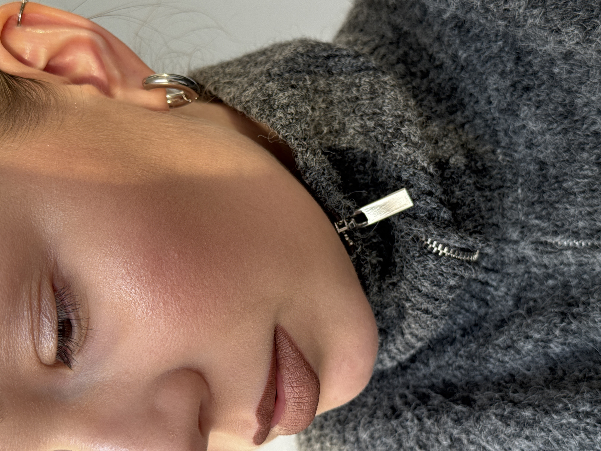 Close-up portrait of a fair-skinned person with brown lips and a piercing in the ear. The person is wearing a grey knit sweater with a visible zipper. Location unknown.