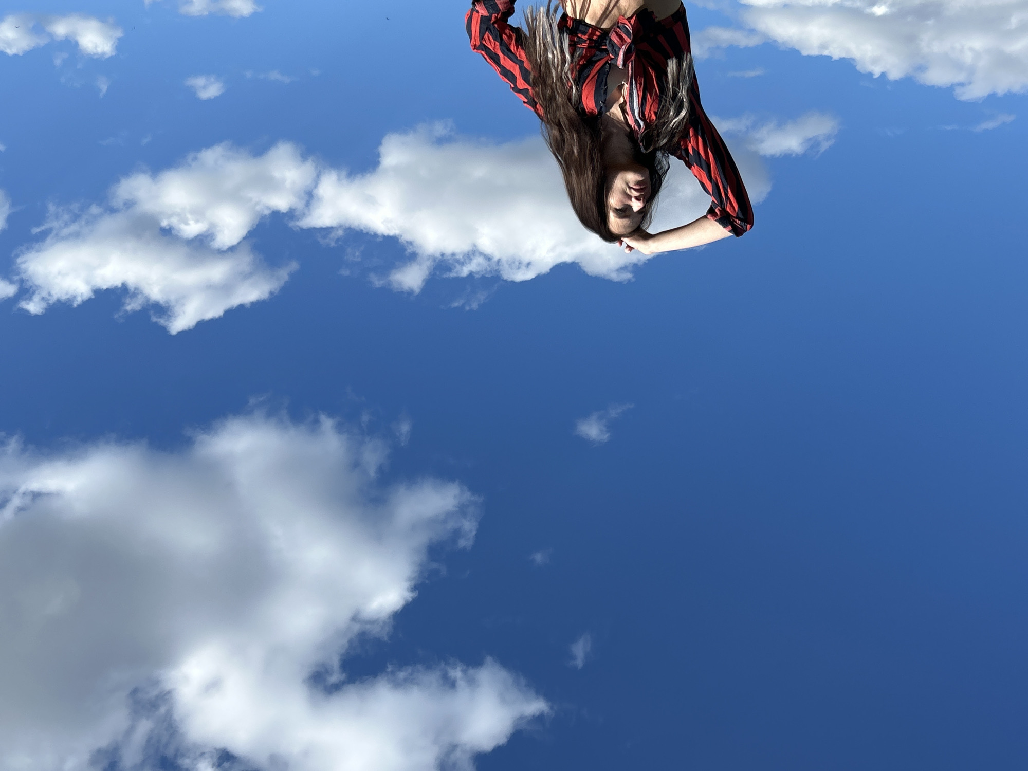 A woman is seen against a blue sky dotted with white clouds. She is wearing a red and black striped shirt tied at the waist. Her long, dark hair falls downwards. The image is rotated 180 degrees. No information suggests this photo was taken in Rusalevka, Russia.