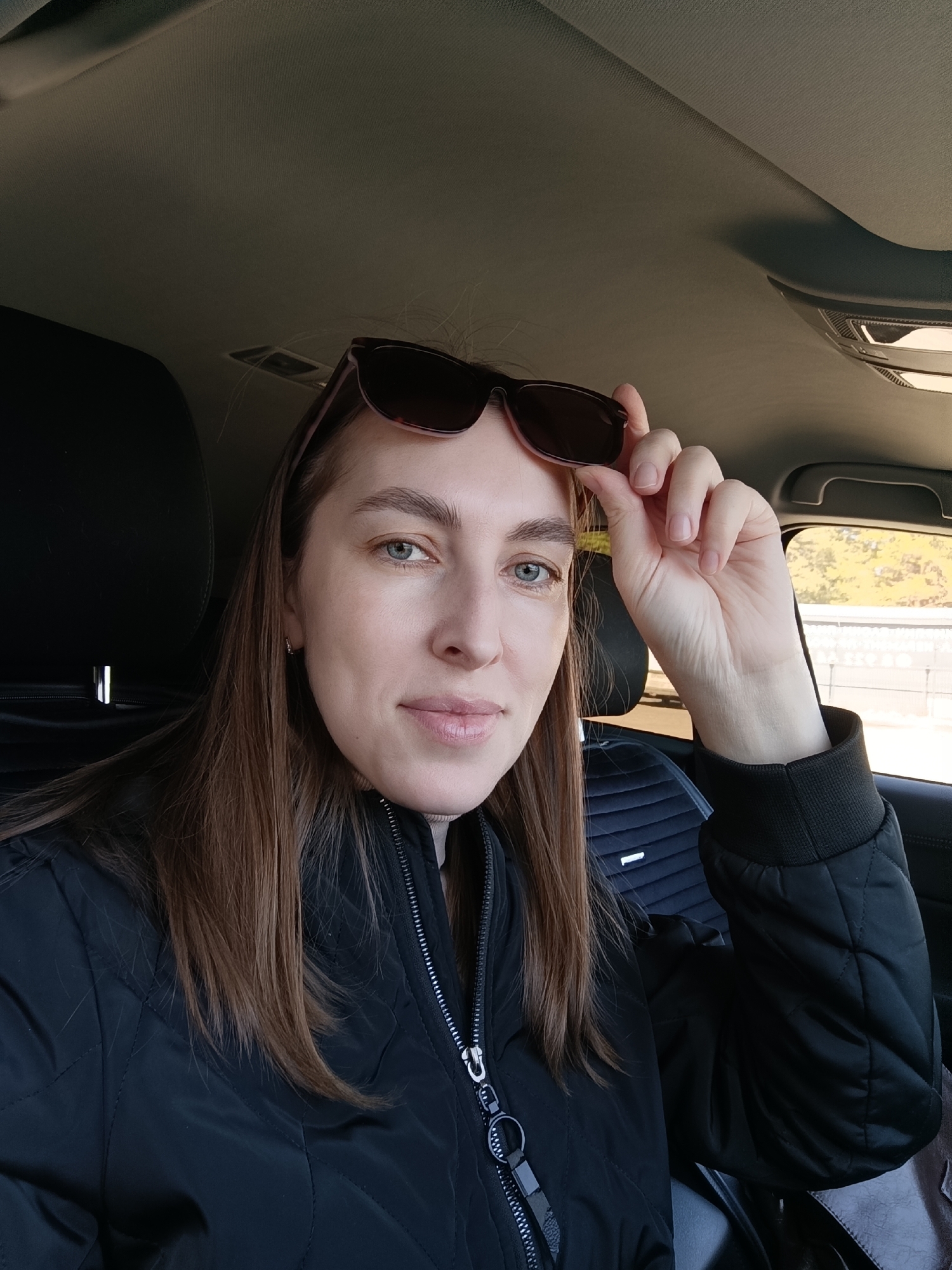 A medium shot of a Caucasian woman with long brown hair sitting in a car. She is wearing a black jacket and holding sunglasses above her forehead. The interior of the car is visible, including the seats and dashboard. Outside the car window, trees and a wall can be seen. The photo was likely taken in the city of Chaikovsky, Russia.