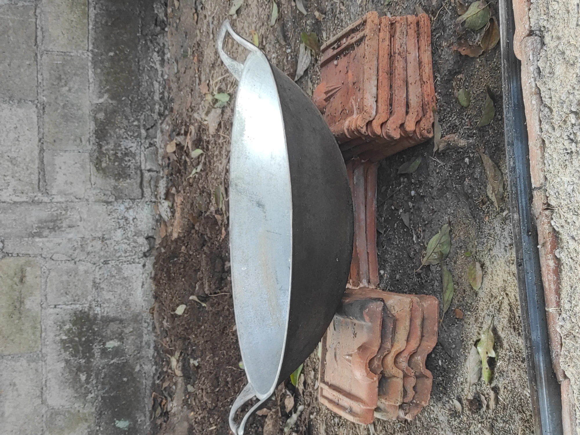 An outdoor scene shows a wok, made of metal, resting on a base constructed from stacked terracotta roof tiles. The wok is positioned centrally, supported by two stacks of tiles. The background consists of a rough, gray brick wall and dark soil, with some scattered leaves on the ground. The overall impression is of a makeshift cooking setup in a rudimentary outdoor area, possibly in Sumedang, Indonesia, given the commonality of such materials and settings.