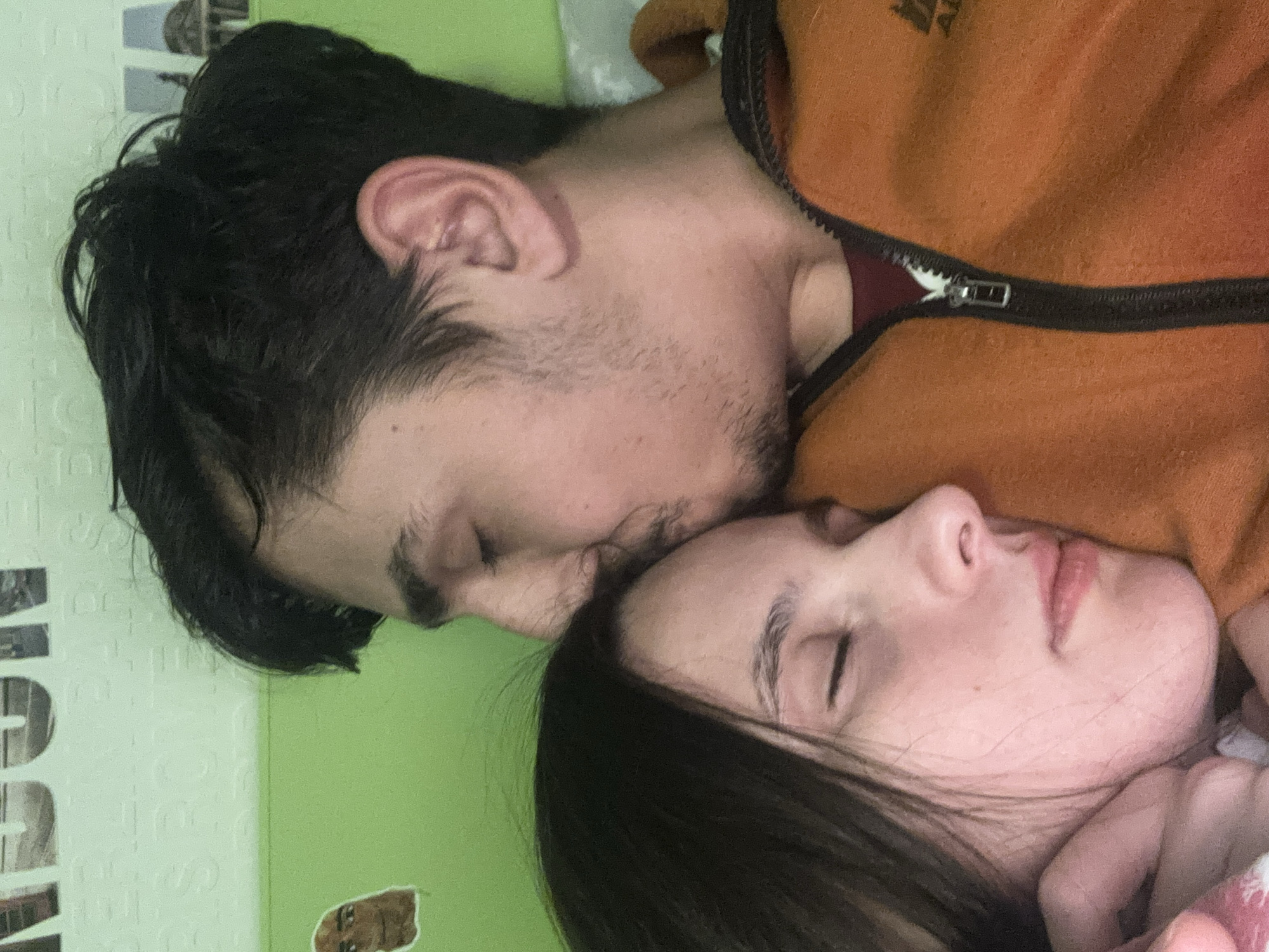 A close-up, overhead shot depicts two people sleeping, with the man lying on his side facing left and the woman facing right. The man has dark hair and stubble and is wearing an orange zippered jacket. The woman has dark hair and is wearing a light-colored garment. The setting appears to be indoors on a green surface with faint patterns, possibly a bed covering. There is no indication that the photograph was taken in Veliko Tarnovo, Bulgaria.