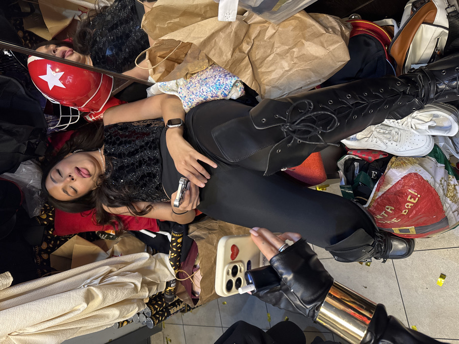 A woman in black leggings and lace-up boots lies amid bags, clothing, and accessories. She wears a sequined top and a watch. A red helmet with a white star sits nearby. Another woman, wearing a black leather glove with a gold band, holds a phone with a red heart sticker. The background is cluttered with items, including a Santa-themed bag, white sneakers, and brown paper bags.