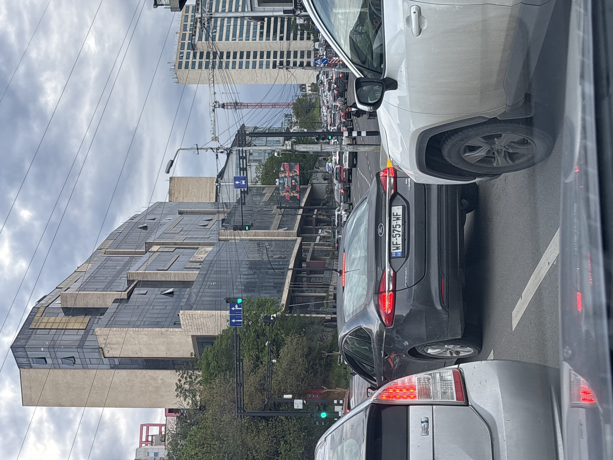 The image shows a street in Tbilisi, Georgia, with heavy traffic. A modern building with a glass facade is on the left. Overhead power lines are visible. In the distance is a building under construction. Several cars are visible in the foreground, including a grey Ford Fusion with a Georgian license plate reading "WF-575-WF" and a white SUV. The traffic lights are green. A large advertising billboard is visible between buildings. The sky is overcast.