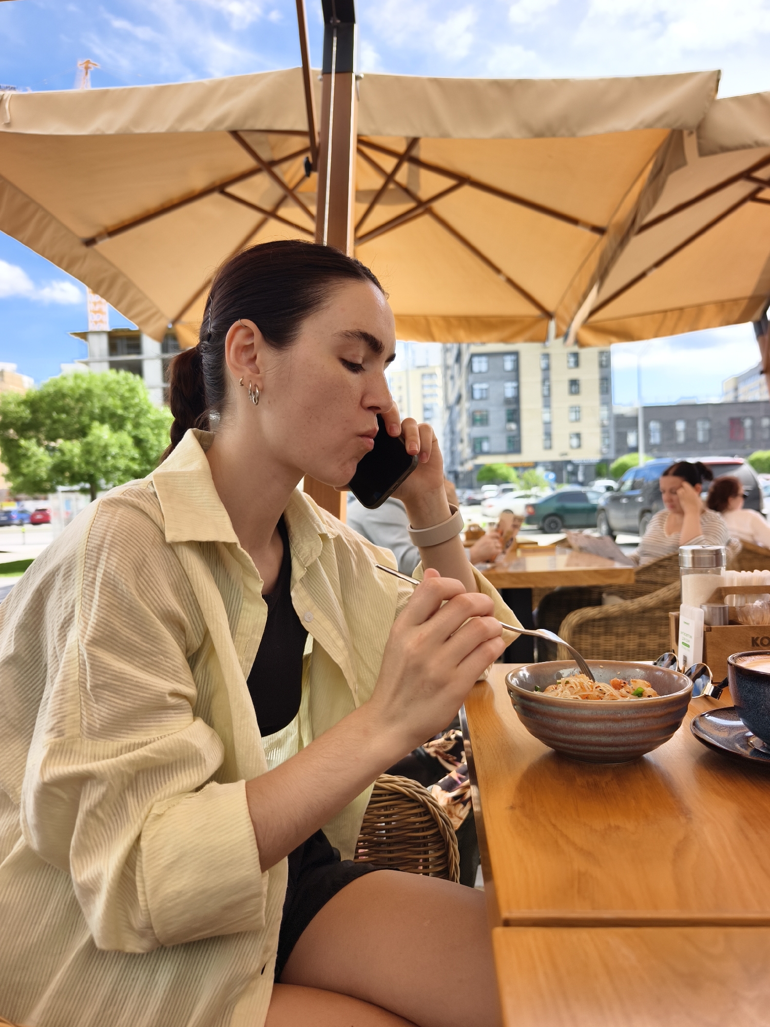 The image captures a young woman dining at an outdoor cafe in Tyumen, Russia, on what appears to be a bright, cloudy day. She is seated at a wooden table, wearing a light yellow striped button-up shirt over a black top, with dark shorts. Her dark hair is tied back in a ponytail, and she has several small hoop earrings in her left ear. She is simultaneously eating a noodle dish (possibly with shrimp and vegetables) from a brown ceramic bowl with a fork and talking on a black cell phone held to her right ear.
On the table near her bowl are sunglasses, and a cup with a saucer, likely containing a hot beverage. A wooden caddy holding a sugar dispenser and salt/pepper shakers is also present, with the text "KOR" visible on its side. She is seated in a wicker chair.
Above the cafe area, large beige umbrellas provide shade. In the background, other patrons are visible at nearby tables. Beyond the cafe's perimeter, an urban street scene unfolds with cars, various buildings (some appearing residential or commercial), and a construction crane in the distance, indicating an active city environment. Trees line parts of the street. The overall setting is a casual outdoor dining experience in an urban landscape.
