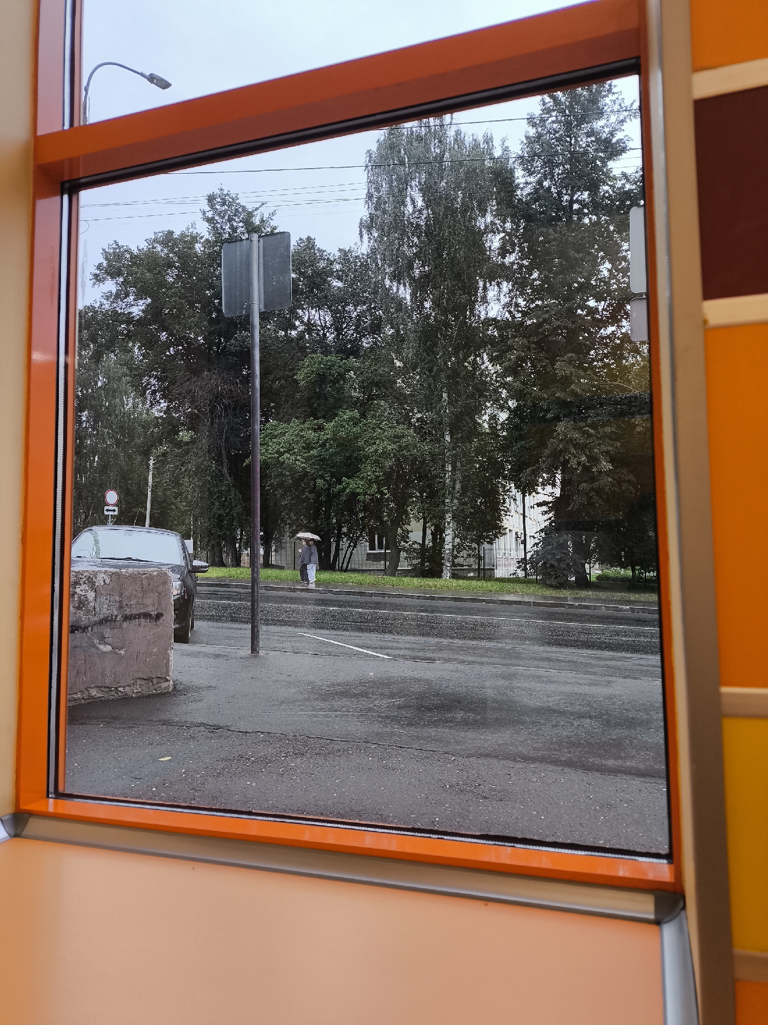 The image shows a view through a window framed in orange, looking out onto a street scene. The sky is overcast, suggesting a cloudy or rainy day. On the left, a dark-colored car is parked on the side of the road, with a concrete barrier in the foreground. A street sign is visible on a pole. Further down the street, two individuals are walking, one holding an umbrella. Trees line the street, with a building partially visible behind them. The road appears wet, indicating recent rain. This scene is consistent with a typical street view in Izhevsk, Russia, given the architectural style and vegetation often found in the city.
