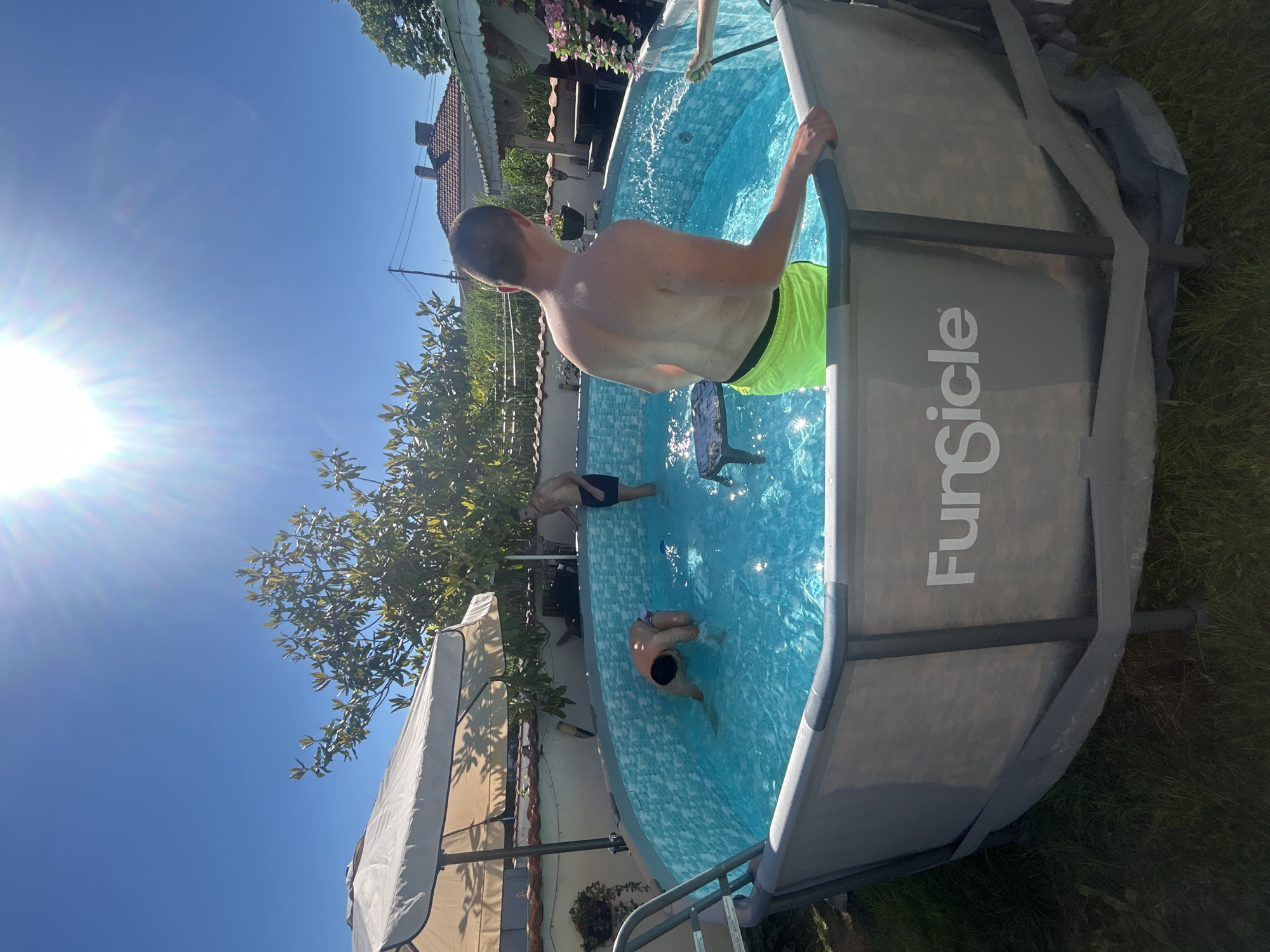 The image captures a sunny day at a backyard pool. The pool is an above-ground, circular model with a gray exterior marked with the word "FunSicle." Inside the pool, several people are swimming. One individual, a man wearing neon green swim trunks, stands near the pool's edge, holding onto the side. In the background, a white gazebo and various buildings can be seen, suggesting a residential area. The location is identified as Elhovo, Bulgaria. A tree and other vegetation are also visible.