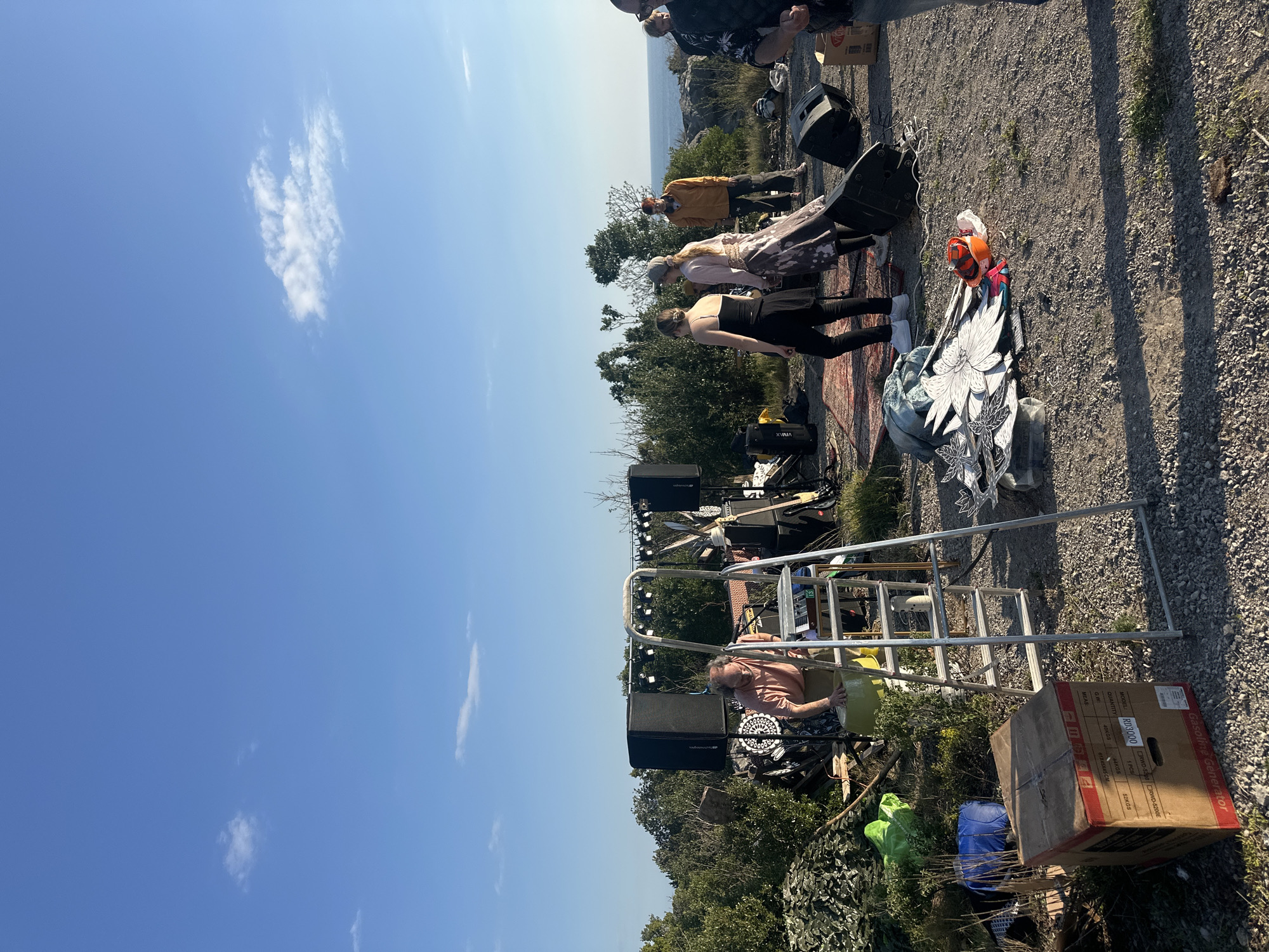 The image depicts a gathering of people outdoors, possibly in the vicinity of Milovići, Montenegro, given the potential coastal view in the background. Several individuals stand in a line, appearing to be engaged in a dance or performance. A woman is seated, possibly managing sound or equipment. A setup of speakers, lighting, a ladder, and other gear suggests a planned event or performance. A cardboard box labeled "Gasoline Generator RD3000" is visible, indicating a power source. The ground is gravelly and uneven, and vegetation is present. The sky is blue with a few clouds.
