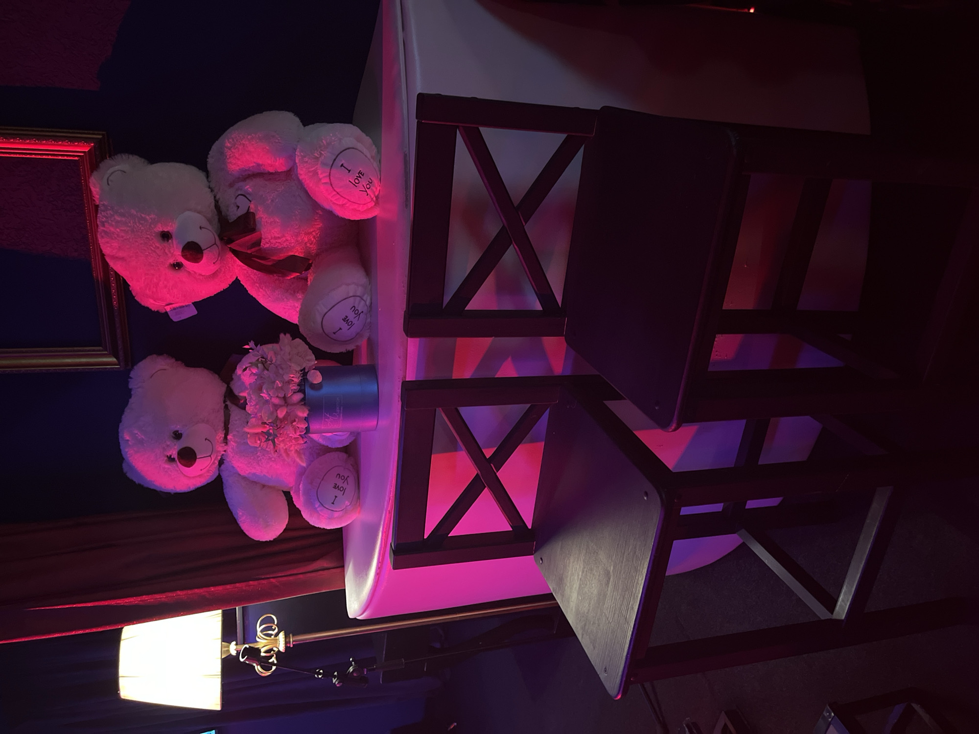 The image depicts a dimly lit room with two large white teddy bears sitting on a white counter. The teddy bears have red bows and "I love you" embroidered on their paws. In front of the counter are two dark wooden bar stools. A lamp with a white shade stands to the left of the scene. The room is illuminated by pink and red lighting, creating a dramatic atmosphere. The location is identified as Kazan, Russia. There are no people present in the image. The scene appears to be set up for a romantic or celebratory occasion.