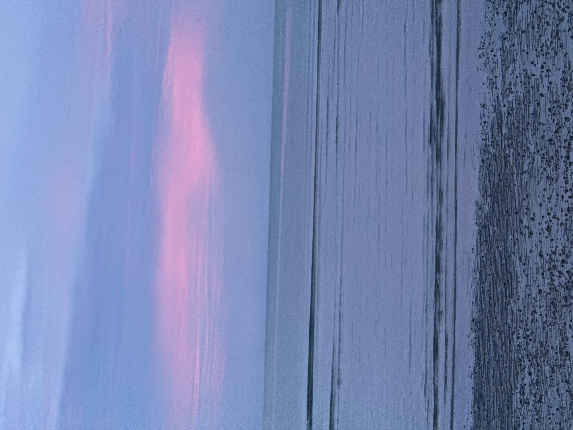 The image depicts a serene coastal scene at what appears to be twilight or dawn. The sky is a soft gradient of blues and purples, with a prominent band of pink clouds stretching horizontally across the upper portion. Below the sky, the calm sea stretches to the horizon, reflecting the muted colors of the sky with faint horizontal lines indicating gentle ripples or wave patterns. The foreground shows a sandy beach with a textured surface, possibly due to receding tides, dotted with small, dark particles. The overall impression is one of tranquility and natural beauty. This scene is consistent with the beaches found in Hua Hin City Municipality, Thailand, known for its picturesque coastline and stunning sunsets.