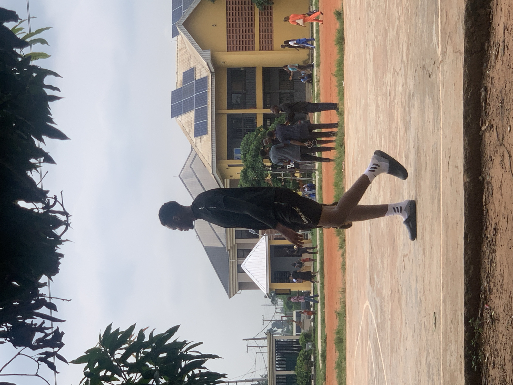 The image depicts an outdoor scene, likely a sports field or court, with a building in the background. Several people are visible, engaged in what appears to be a sports activity or training. The primary subject in the foreground is a young man in athletic attire, a black t-shirt and shorts, with white socks and dark athletic shoes. He is in motion, his body positioned as if mid-stride or jump, with one leg extended and the other bent.
In the background, a yellow building with solar panels on its roof is prominent. A group of other individuals can be seen near the building, some in athletic wear, suggesting a sporting event or practice. The ground is a mix of reddish-brown dirt and a lighter colored surface, possibly a court. Lush green foliage is visible on the left and bottom edges of the image, framing the scene. The sky appears overcast, suggesting a cloudy day. The time of day is likely daytime, given the lighting conditions. There is no discernible text in the image. The overall atmosphere seems to be one of athletic activity and training in a somewhat casual, outdoor setting.