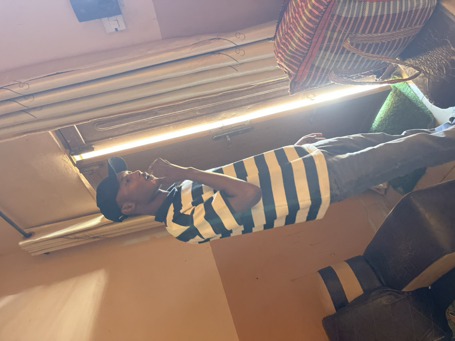 An indoor scene depicts a male individual standing near a brightly lit doorway in a room with light brown walls. The individual, positioned slightly right of center, faces right in profile, wearing a black baseball cap, a navy blue and white horizontal striped short-sleeve polo shirt, and dark trousers. His right hand is raised towards his mouth or chin. Strong sunlight emanates from the open doorway behind him, illuminating the subject's right side and creating a bright vertical strip in the center background. Light-colored curtains are drawn to either side of the doorway. To the far left, a section of a dark brown armchair or sofa with light accents is visible. To the far right, a large tote bag with red, white, and possibly green or blue horizontal stripes and woven handles rests on a dark surface. A small green mat is on the floor directly behind the standing individual. Above the curtains on the right, a dark object, possibly a speaker, is mounted on the wall next to a white electrical outlet. The specific location within Unguwar Gabas, Nigeria, is not discernible from the visual information provided.