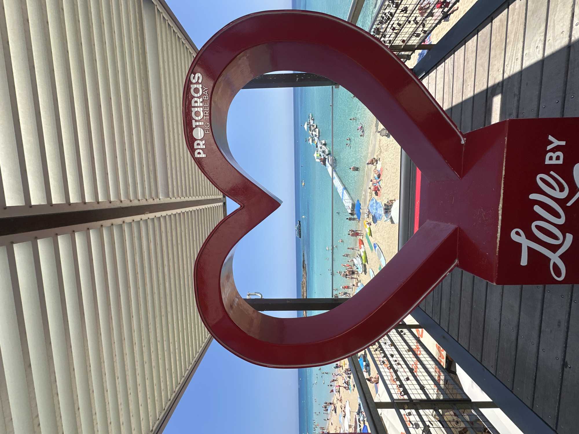 A vibrant red heart-shaped sculpture is positioned in the foreground, framing a picturesque view of Fig Tree Bay in Protaras, Cyprus. The sculpture, adorned with the words "PROTARAS" and "FIG TREE BAY" in white lettering at the top, and "Love BY" in cursive white lettering at the bottom, stands on a wooden deck. Beyond the heart, the turquoise waters of the bay teem with activity. Numerous people are visible on the sandy beach and swimming in the sea. A long, white jetty extends into the water, lined with several boats. The sky is a clear, brilliant blue, and the scene is bathed in bright sunlight, indicating a warm, sunny day. The overall impression is one of a popular tourist destination, offering relaxation and recreation.
