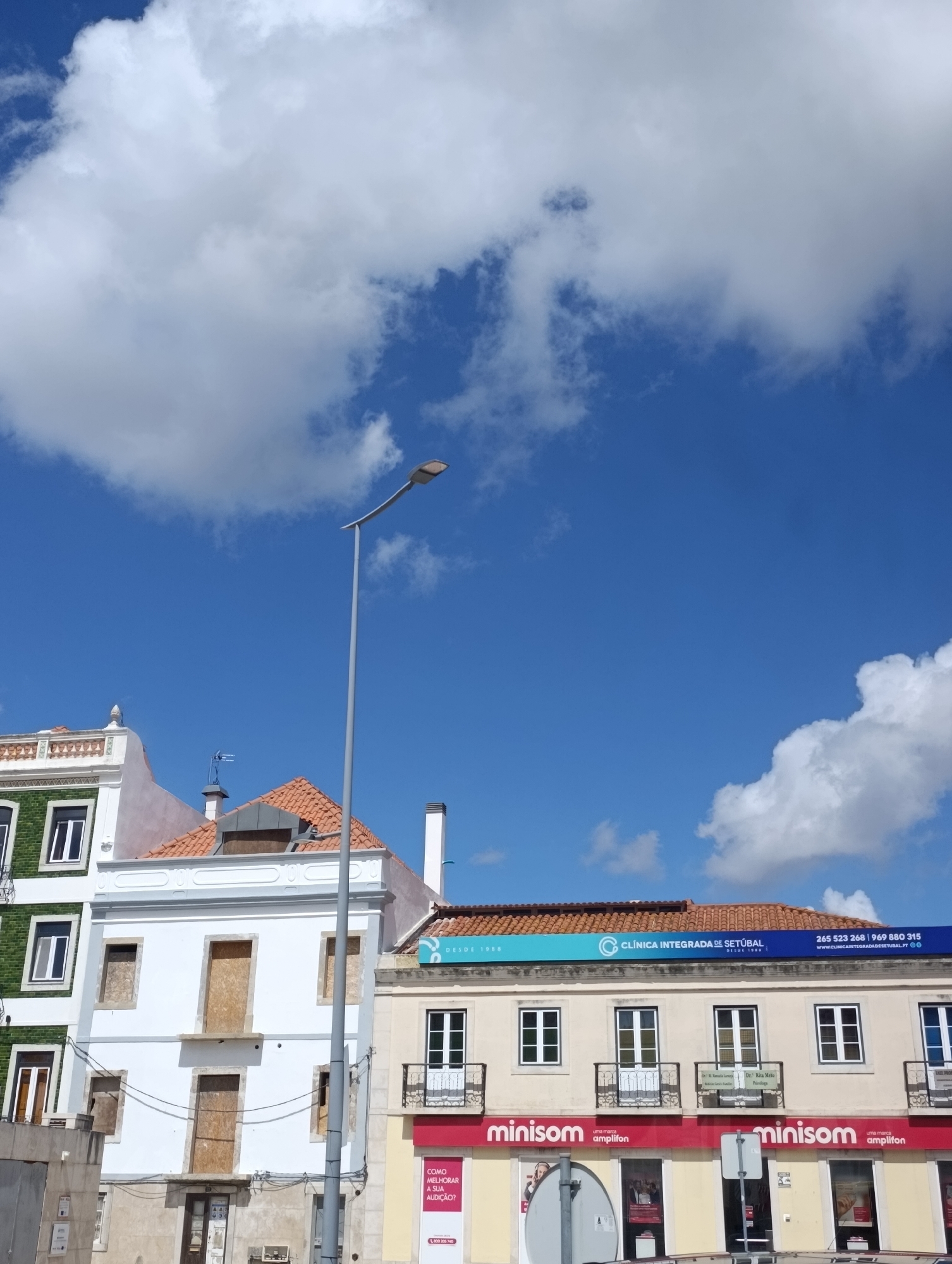 The image captures a street-level view of buildings in Setúbal, Portugal, under a bright blue sky dotted with white clouds. The architecture features a mix of styles, with the foreground dominated by a white building with boarded-up windows, suggesting it might be under renovation or disuse. To its left, a building with green tiled walls and white-framed windows stands, also appearing to be residential or commercial.
Above these, a prominent light pole with a curved arm extends into the frame from the right side. Further back, a building with an orange-tiled roof displays a blue sign that reads "CLÍNICA INTEGRADA DE SETÚBAL DESDE 1990" along with a phone number and website, indicating a medical clinic. Below this sign, the ground floor of the building hosts a "minisom" store, identifiable by its red awning and signage.
The overall scene presents a typical urban streetscape in Portugal, with a blend of older and more contemporary architectural elements. No people are clearly visible in the image, and the focus is on the buildings and the sky.