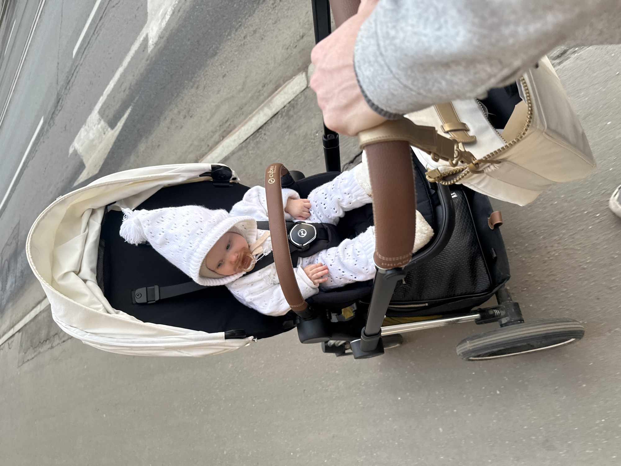 A first-person perspective image depicts an infant seated in a white and black Cybex stroller being pushed along a paved surface in Moscow, Russia. The infant, wearing a white knitted outfit including a matching hat with a pompom, has a brown pacifier in its mouth and brown residue around its lips. The child is secured by black harness straps. A human hand, wearing a grey long-sleeved garment, is visible in the foreground, gripping the stroller's brown leatherette handle. The hand also holds a light beige handbag with a gold zipper and chain strap. To the right and in the background, a grey asphalt road is visible, featuring white painted markings, including a double solid line and diagonal chevron patterns, with visible tire tracks. The stroller's black wheels, which have white rims, are visible. The environment is outdoor, suggesting daylight.