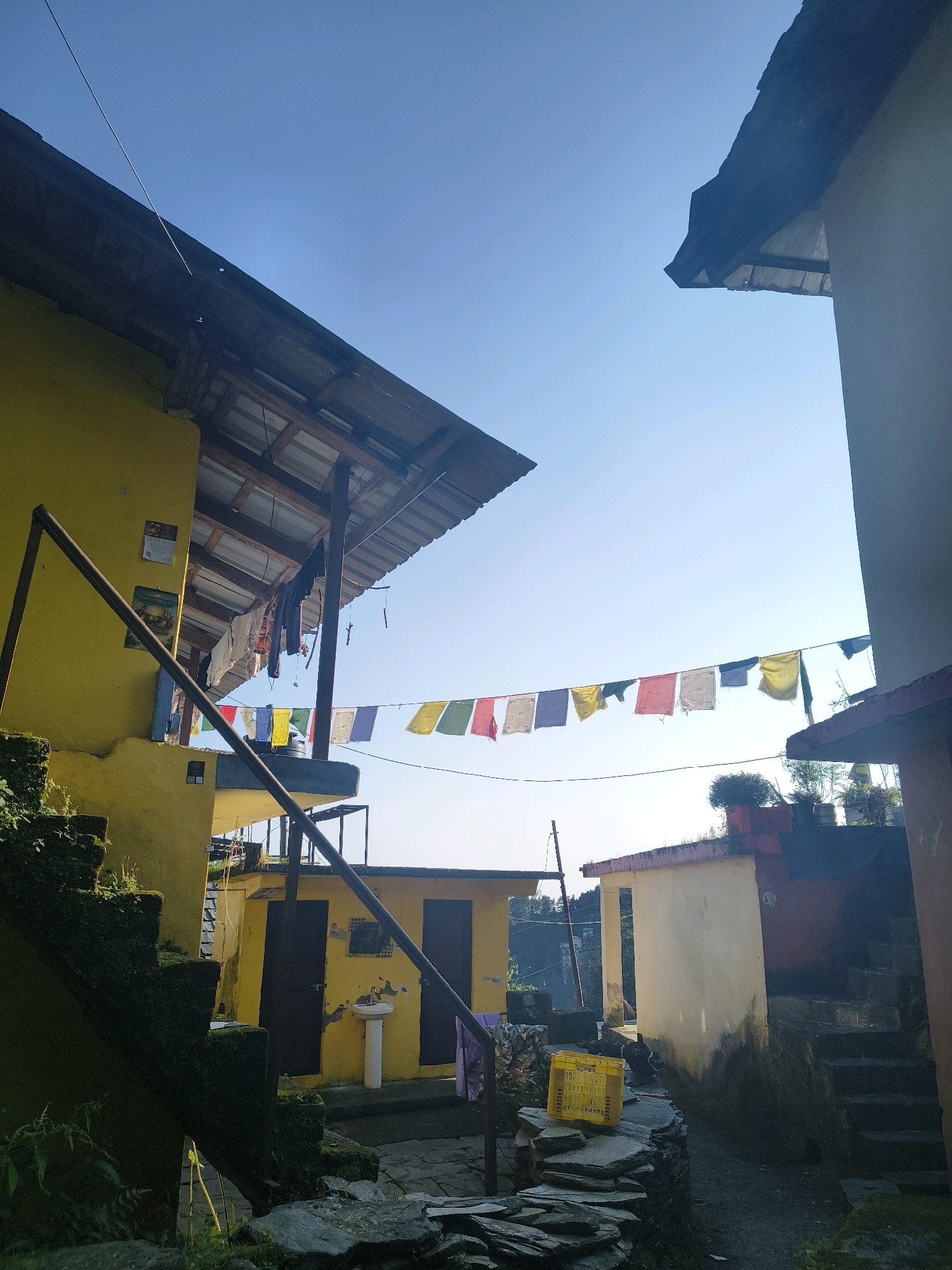 The image depicts an outdoor scene, likely in Haini, India, characterized by bright sunlight and a clear blue sky. In the foreground, a stone pathway leads through a narrow alleyway between two buildings. Moss-covered stone steps are visible on the left, leading up to a yellow building with a corrugated metal roof supported by wooden beams. Laundry hangs on a clothesline strung across the balcony of this building.
Further into the scene, prayer flags in various colors are strung horizontally across the pathway, adding a vibrant element to the otherwise muted tones of the architecture. These flags are a common sight in Himalayan regions, often associated with Buddhist monasteries and spiritual sites.
The buildings themselves appear to be multi-story structures with weathered facades, suggesting a rural or traditional setting. One building on the right is painted in a light yellow, with a reddish trim along the roofline and stone steps leading up to its entrance. Another structure, partially visible on the far right, is a lighter color and features a darker roof.
The overall impression is one of a tranquil, lived-in environment, possibly a village or a guesthouse, where daily life unfolds amidst the backdrop of natural beauty and cultural traditions. The clear sky and bright sunlight suggest it is a pleasant day, ideal for outdoor activities.