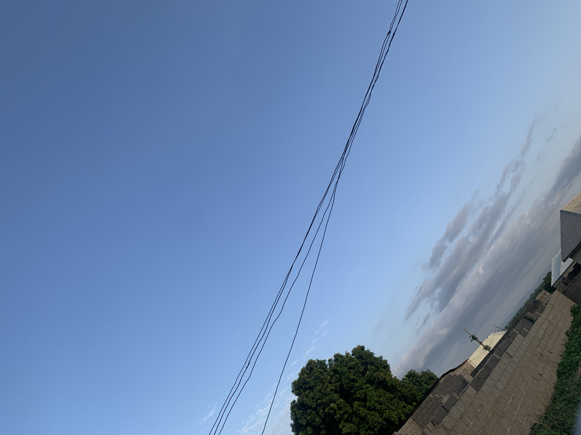 The image captures a daytime scene in Keffi, Nigeria. The sky is predominantly blue with some thin clouds near the horizon. Three electrical wires run diagonally across the frame. In the lower right, there's a tree with dense green foliage. Adjacent to the tree is a building under construction, made of gray cement blocks. A roof is visible on the building. Some vegetation can be seen alongside the partially built structure.