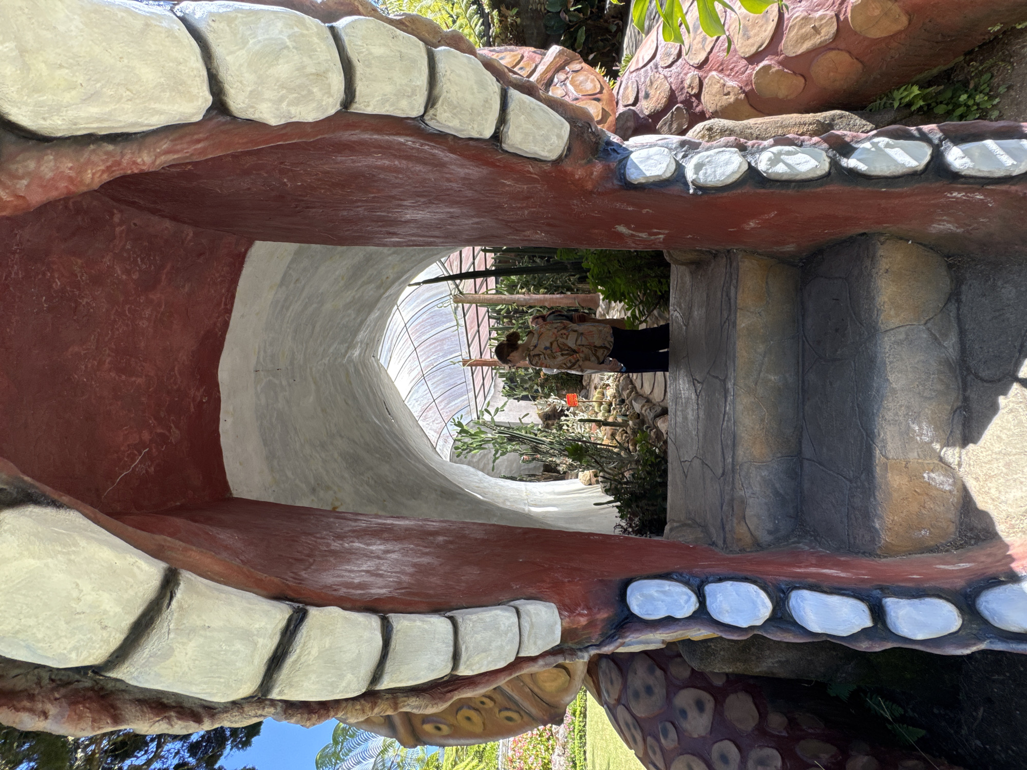 This image depicts the entrance to a botanical garden in Baturiti, Indonesia, designed to resemble the gaping mouth of a monster or creature. The entrance is framed by large, textured elements painted red and white, with oversized, white, tooth-like structures lining the opening. Stone steps lead up into the mouth, which serves as a tunnel. Inside the tunnel, a person wearing a patterned shirt and dark pants stands looking towards the interior of the garden. The garden itself is visible beyond the tunnel, featuring a variety of plants, including cacti and other greenery, under a translucent, arched roof structure. The overall scene suggests an attraction within a tourist destination, likely a garden or park.