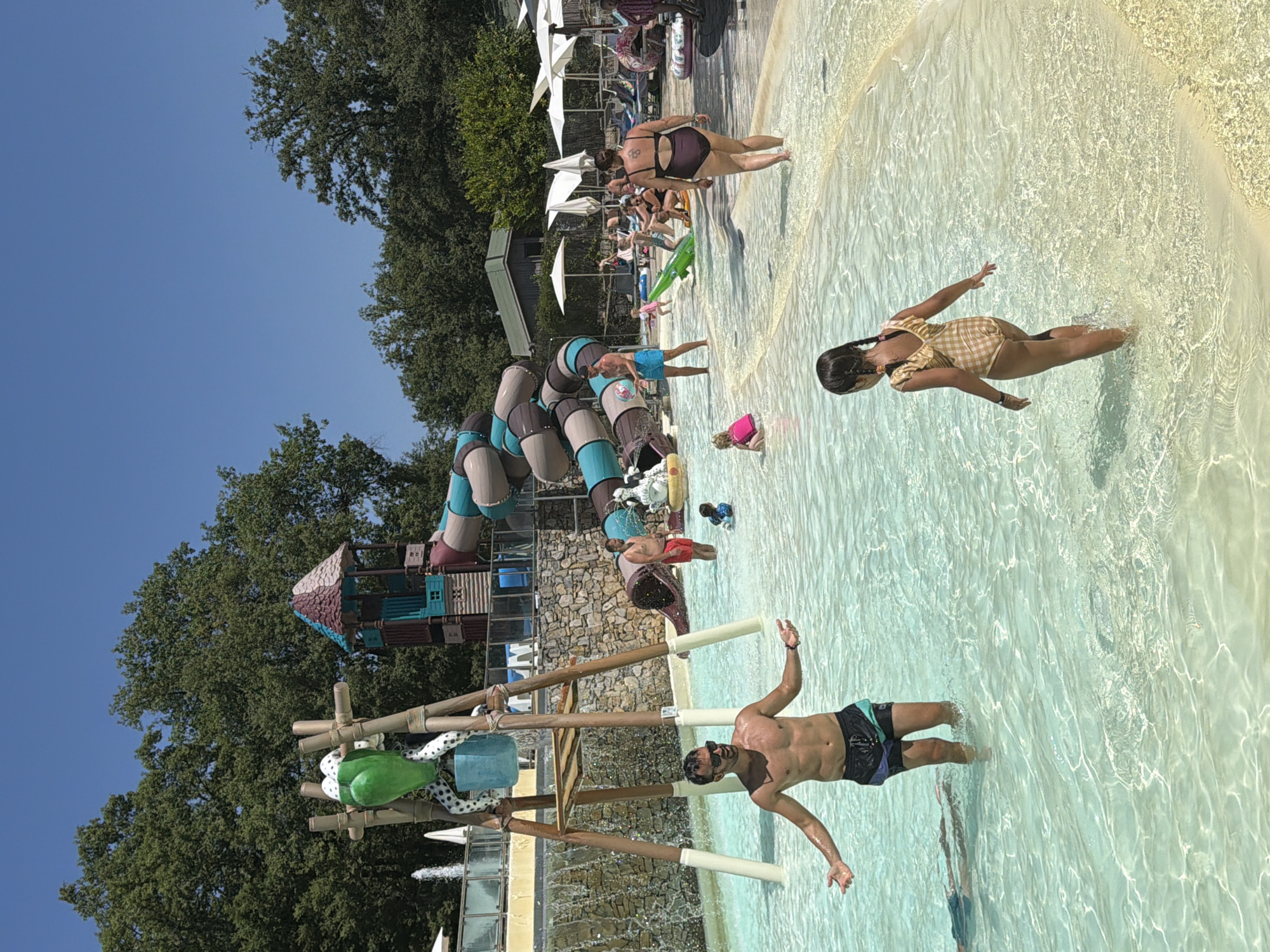 This image depicts a sunny day at a water park, likely within the Tuscan region of Italy, and specifically located in Cavriglia. In the foreground, a man stands in shallow, clear water, gesturing with his hands. Behind him, a young girl in a checkered swimsuit walks away from the camera, her hair in braids. Further back, several other individuals are visible, including a man in red swim trunks near a water slide, and a woman in a dark swimsuit walking away. The water park features several attractions, including a large, winding water slide structure with a playful design. A decorative element resembling a green frog with polka-dot legs is attached to a wooden structure. In the background, trees and umbrellas suggest a recreational outdoor setting. The water is shallow and sandy at the edges, with clear, illuminated water in the main area.