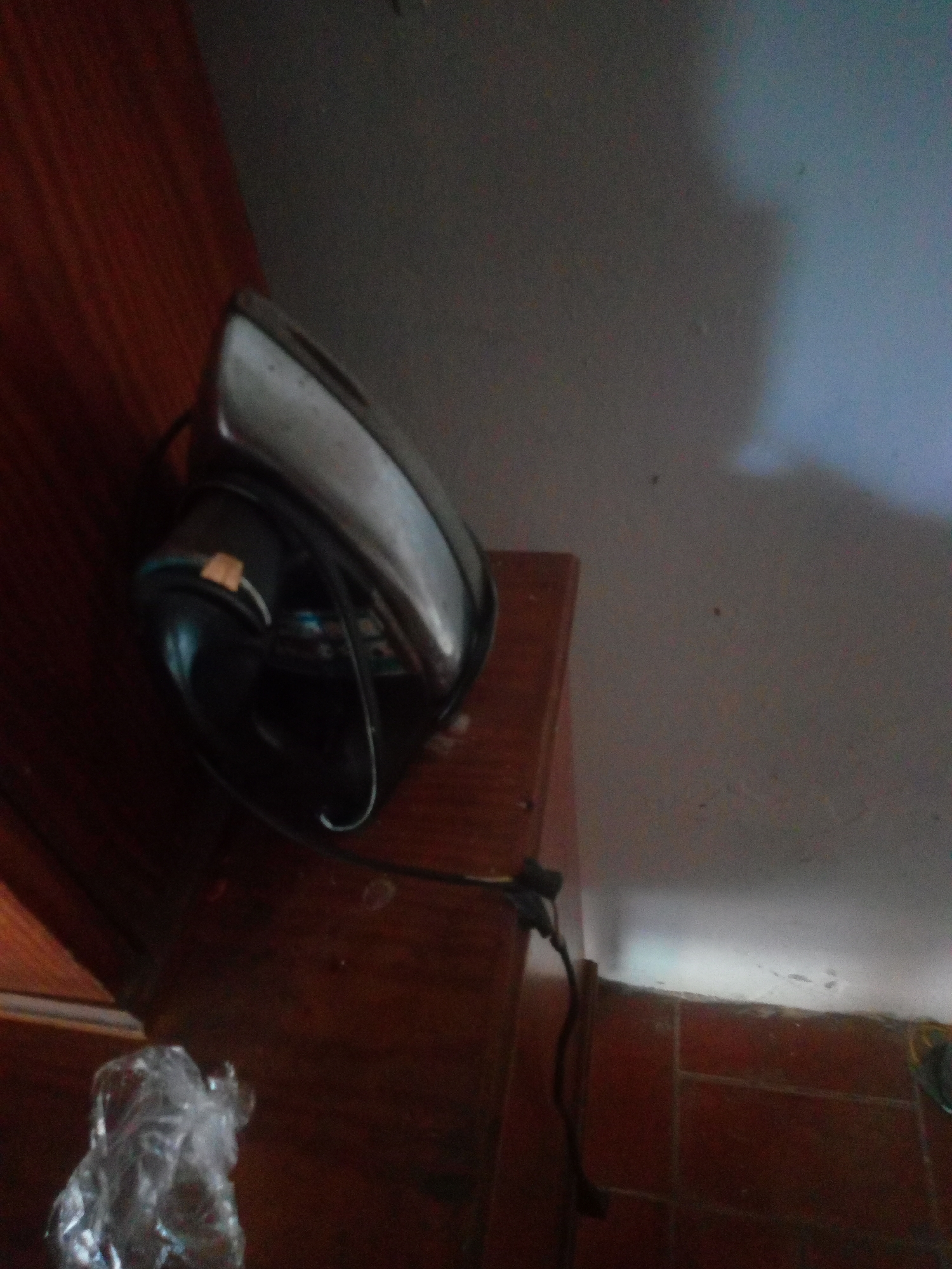 An iron is resting on a wooden surface. The iron is silver and black with a black cord. The wooden surface appears to be a table or dresser. To the left of the iron, a small plastic bag is visible. The background is a wall, with a shadow cast on it. The scene is indoors, likely a room in a house in La Tagua, Venezuela.