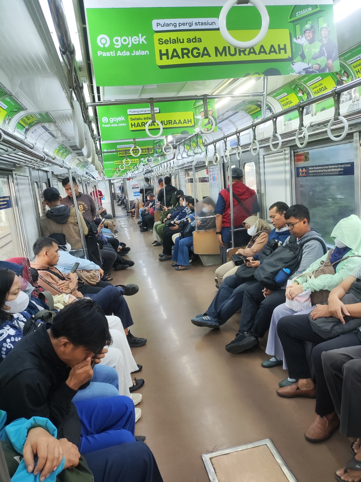 The image depicts the interior of a commuter train carriage, likely in Tambun, Indonesia, based on the context provided. Passengers are seated and standing within the train. Advertisements, primarily in green and white, are visible along the top of the carriage, featuring the Gojek logo and promotional messages. Many passengers are wearing face masks. Some individuals are looking at their phones, while others are resting or conversing. The train carriage appears to be well-lit by overhead lights. The floor of the carriage is a light brown color. Various handrails and poles are present for passengers to hold onto.