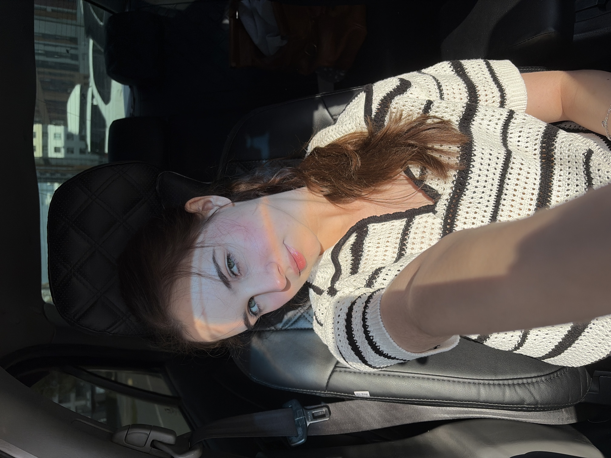 A young woman with dark hair and light skin is lying in a car seat. She is wearing a black and white striped shirt. The car is parked, and buildings are visible through the window. The interior of the car is dark. The scene appears to be naturally lit with sunlight. It is not possible to determine the specific location of the photo from the information provided.