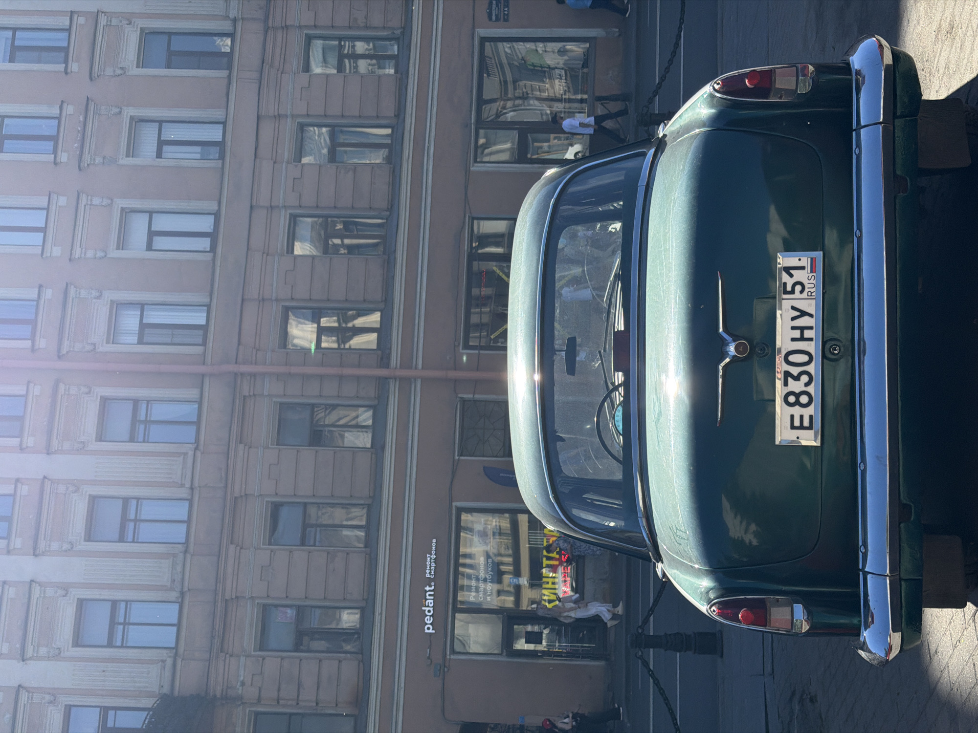 The image displays the rear view of a vintage dark green GAZ-21 Volga sedan parked on a street in Saint Petersburg, Russia. The vehicle's Russian license plate reads E830HY 51 RUS, indicating its registration from the Murmansk Oblast. The car features a prominent chrome bumper and two vertical taillights with red and white segments. The trunk lid has a central winged emblem.
In the background, a multi-story, light pink or salmon-colored building with numerous rectangular windows constitutes the street facade. The upper floors exhibit classical architectural elements, including decorative cornices above windows and pilasters between them. At street level, the building houses commercial establishments. On the left, a storefront with a white sign reads "pedant. ремонт смартфонов" (pedant. smartphone repair). Below it, through the window, additional signage for "РЕМОНТ СМАРТФОНОВ И НОУТБУКОВ" (REPAIR OF SMARTPHONES AND LAPTOPS) is visible, along with partial text like "КИНЕТА" and "VAPE S".
Several individuals are present on the sidewalk. To the left of the car, near the "Pedant" shop, a person in light clothing walks away from the viewer, while another person wearing a red helmet or hat is partially visible further left. To the right of the car, a person in a white t-shirt and dark pants walks towards the right side of the frame. The scene is illuminated by bright sunlight, casting reflections on the car's surface and building windows, and suggesting a daytime setting. Black bollards connected by chains delineate the curb line in the foreground.
