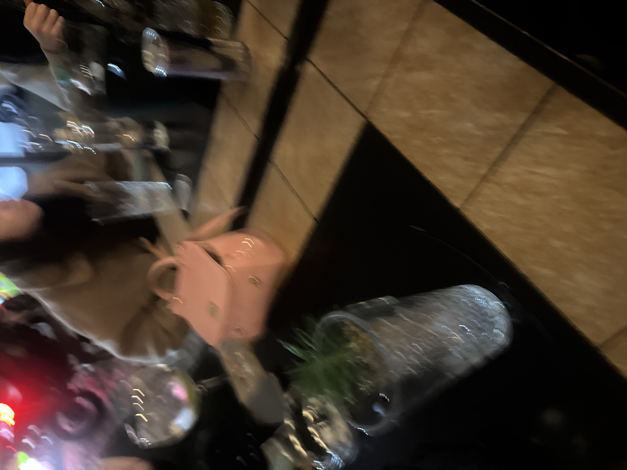 A blurred, high-angle shot depicts a table setting in an unidentified location in Plovdiv, Bulgaria. A person is partially visible on the left, wearing a light brown coat and possibly sitting at the table. A pink purse sits on the table next to the person. There is a clear glass with a small plant inside on the table. Other objects such as glassware are also visible. The table is dark. The surrounding environment appears to be a restaurant or bar, indicated by the table setting and other people in the scene. The background consists of flooring, possibly tile, in shades of brown. Due to the blur, specific details are indistinct.