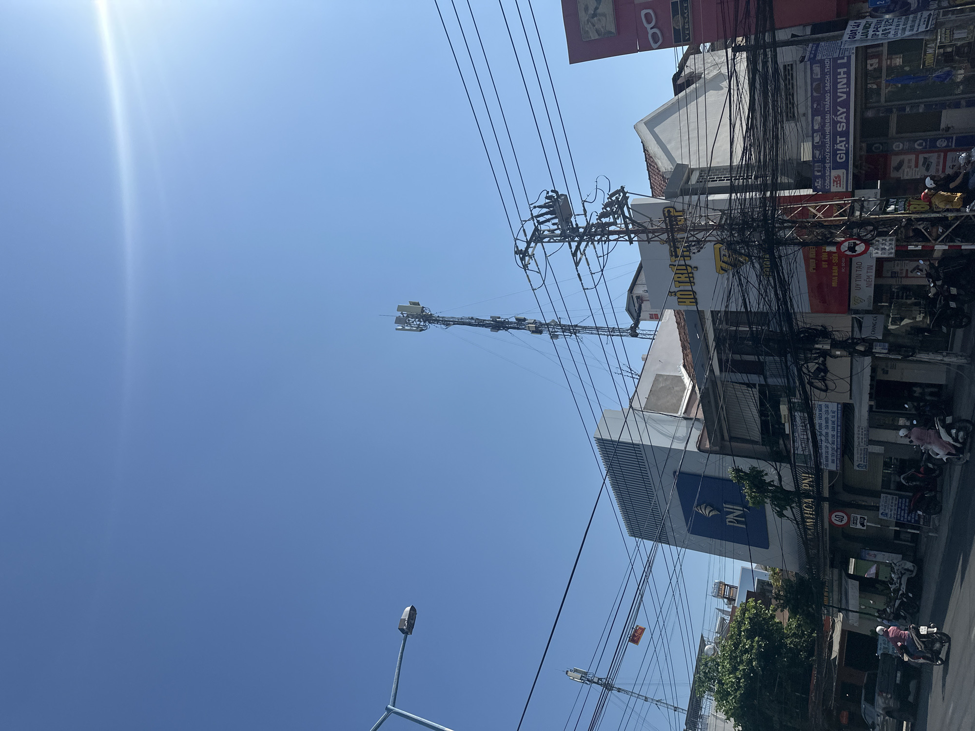 The image depicts a street scene in Bac Nha Trang Ward, Vietnam, characterized by a clear blue sky and an abundance of utility poles and power lines crisscrossing the air. The foreground features a streetlight, while the middle ground is dominated by buildings with commercial signage, including "PNI," "VERSACE," and laundry services ("GIẶT SẤY VỊNH LE"). Several motorcycles are parked or moving along the street. A communications tower stands tall amidst the buildings. Overhead power cables are heavily clustered around the buildings. There are no individuals readily identifiable in the frame.