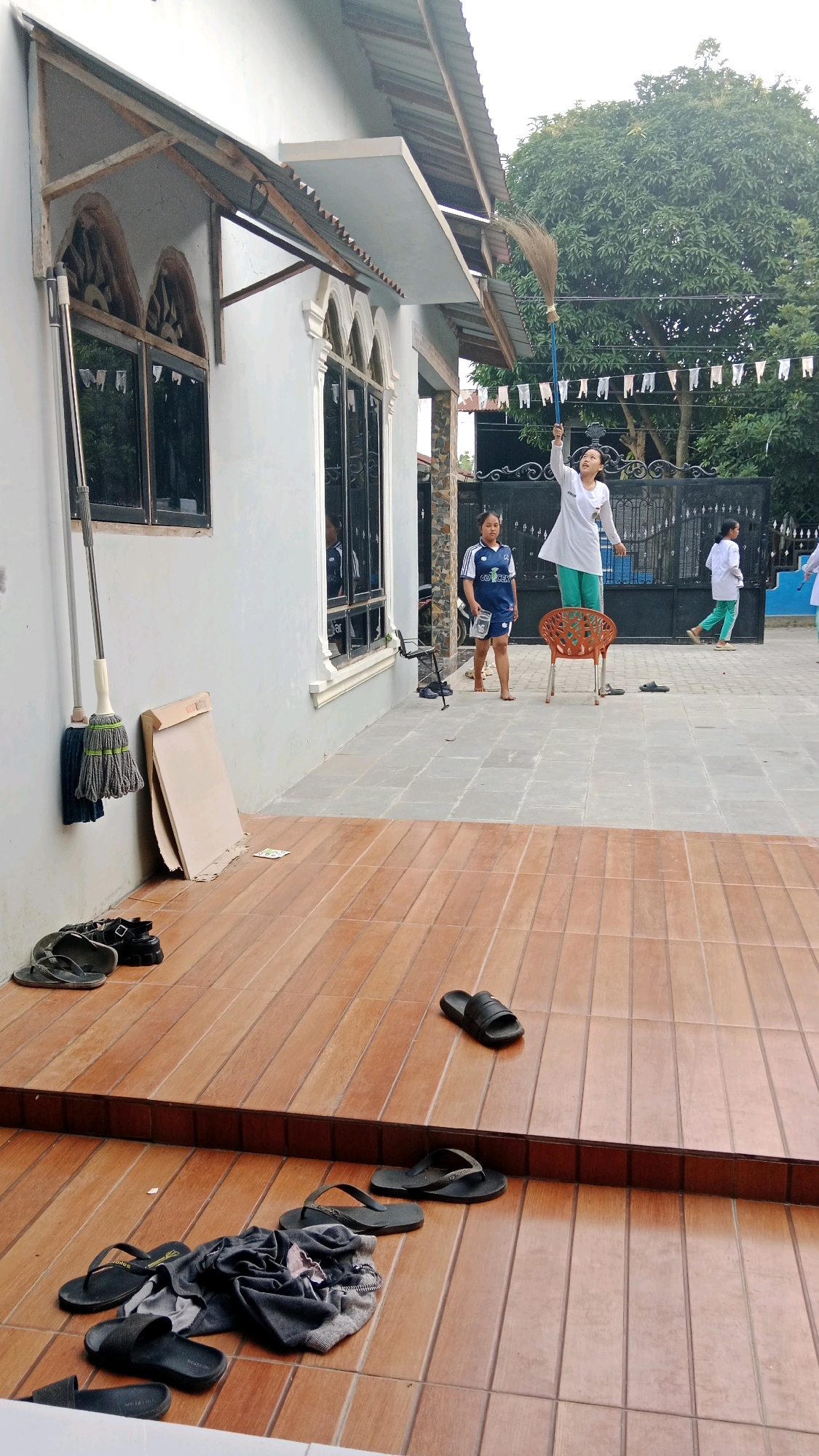 In Medan Senembah, Indonesia, an outdoor scene unfolds. A young female, positioned on an orange plastic chair, is actively sweeping with a broom that extends upwards. To her left, another young female in a blue and white soccer jersey stands observing. Further to the right, a third young female in a white top and green pants is walking away from the camera, while another individual, also in a white top and green pants, is visible in the background. The setting appears to be a residential outdoor area with a light-colored building featuring arched windows and a covered walkway. Paving stones and wooden decking form the ground surface, and various footwear, including several pairs of flip-flops and sandals, are scattered around. A bundle of clothing lies on the wooden deck, and two mops are propped against the building wall. A metal gate and lush green trees are visible in the background, along with decorative white flags strung between poles.