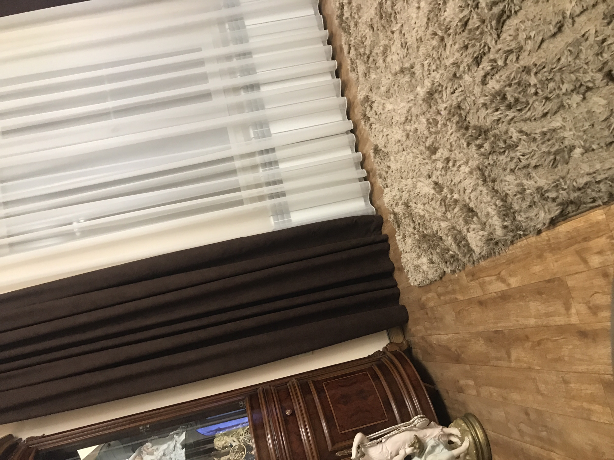 The image displays a room interior, capturing elements such as curtains, a shag rug, hardwood flooring, and a piece of furniture, possibly an antique display cabinet or credenza. The curtains are layered, featuring sheer white horizontal blinds with a darker brown fabric curtain underneath. The shag rug, appearing in beige or light brown, is adjacent to the wooden floor. The furniture piece shows a curved top and decorative details, suggesting an older, possibly European-influenced style. Yerevan, Armenia, is not visibly represented in this image.