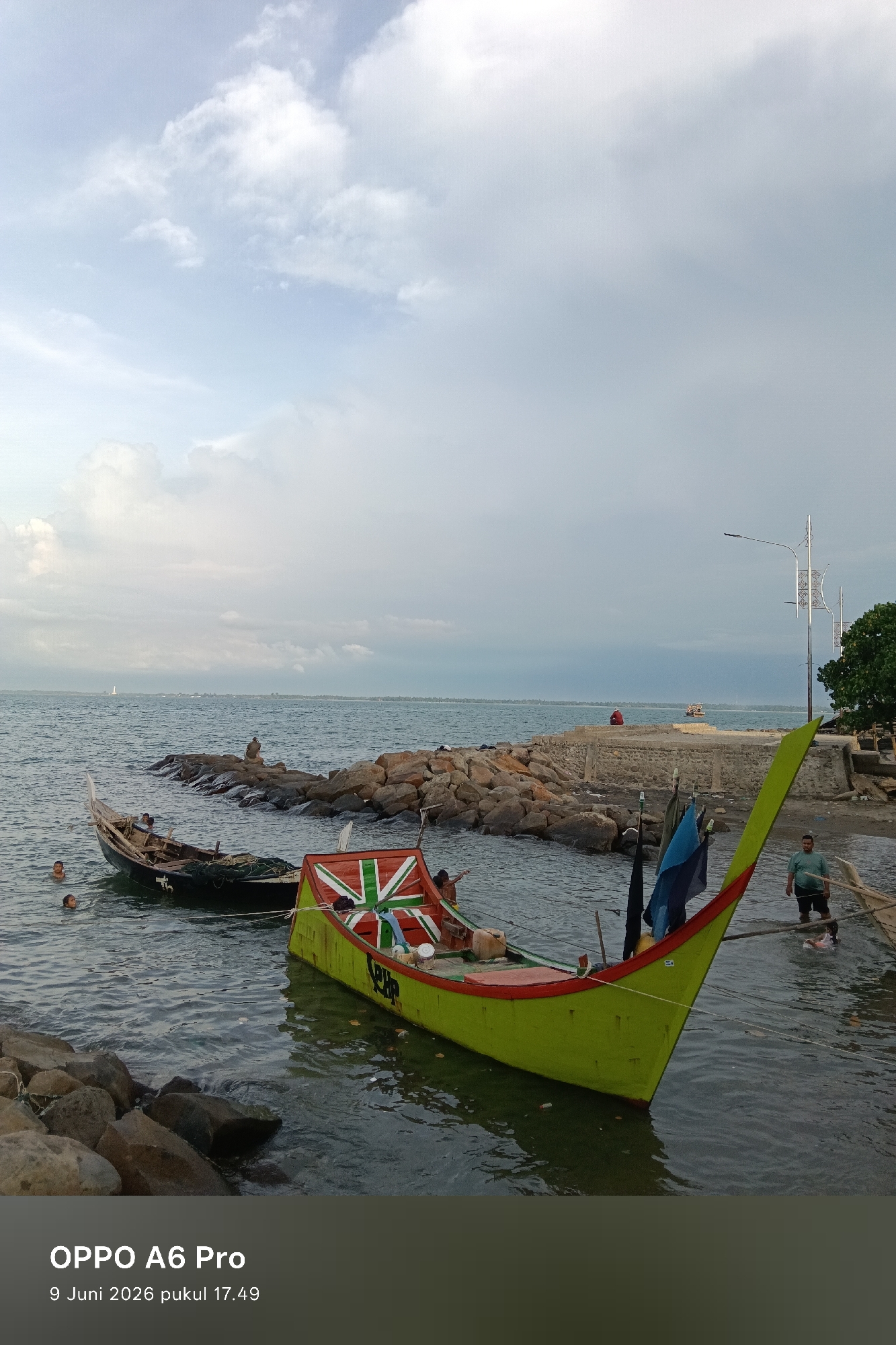 The image depicts a waterfront scene with two boats in the foreground. The boat on the left is partially submerged and appears older, with several people in the water around it. The boat on the right is painted bright green with red trim and features a flag with a Union Jack design. Several people are visible near the boats, some in the water and some on the shore. In the background, there is a rocky breakwater extending into the sea, with more distant land visible on the horizon. The sky is overcast with scattered clouds. The timestamp at the bottom indicates the image was taken in Lhokseumawe, Indonesia, on June 9, 2026, at 5:49 PM.