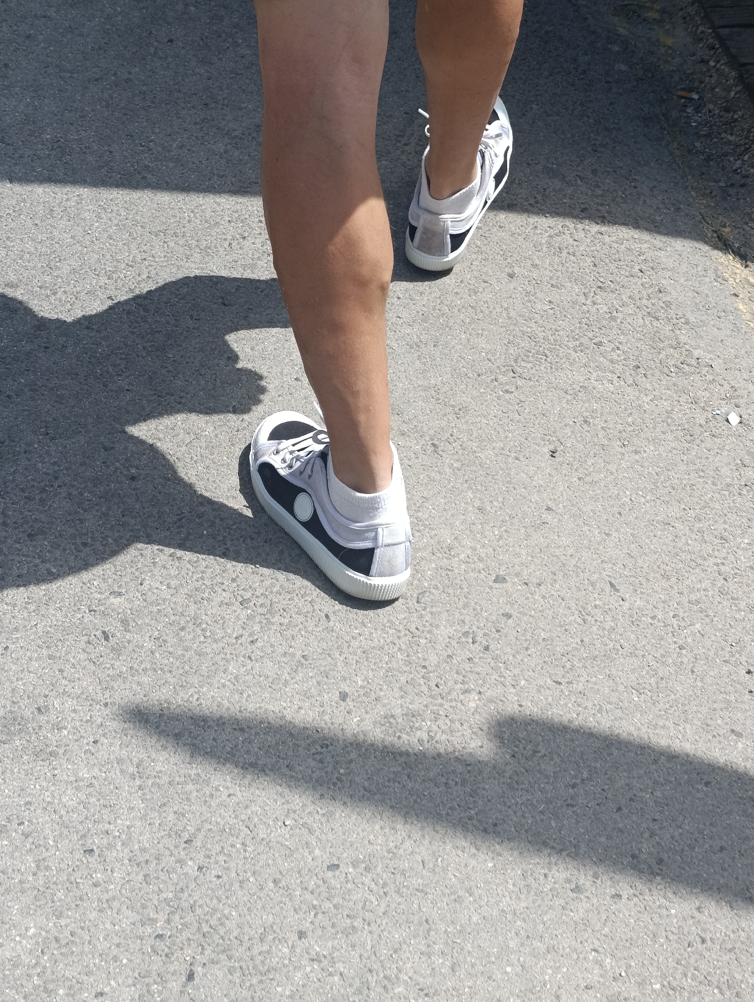 A close-up, eye-level shot shows the lower legs and feet of a person walking on an asphalt surface. The person is wearing white athletic shoes with black accents and a white ankle sock. A distinct shadow is cast on the ground to the left of the person, indicating bright sunlight from the right. The asphalt appears to be a city street or sidewalk, with some small debris visible. The background is out of focus, suggesting a shallow depth of field. The overall impression is a casual, outdoor scene. The location of Setúbal, Portugal, is not directly inferable from the visual elements within the image.