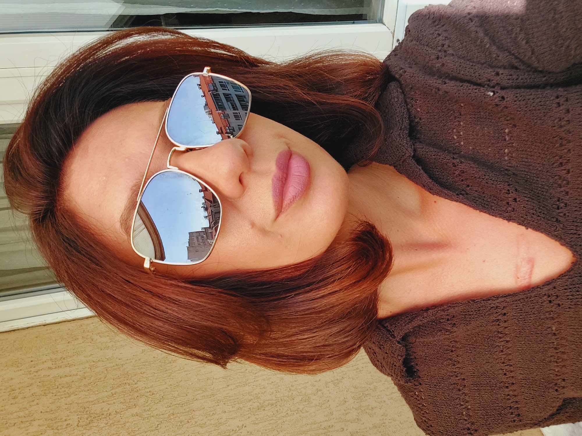 A close-up, eye-level, outdoor, selfie-style photograph shows a fair-skinned woman with shoulder-length, auburn hair wearing aviator-style sunglasses and a brown, knitted, collared shirt. The sunglasses reflect buildings and a blue sky. A scar is visible on the woman's chest. The background consists of a textured, tan wall on the left and a window frame with white blinds on the right. The scene appears to be on a balcony or patio in Sofia, Bulgaria.