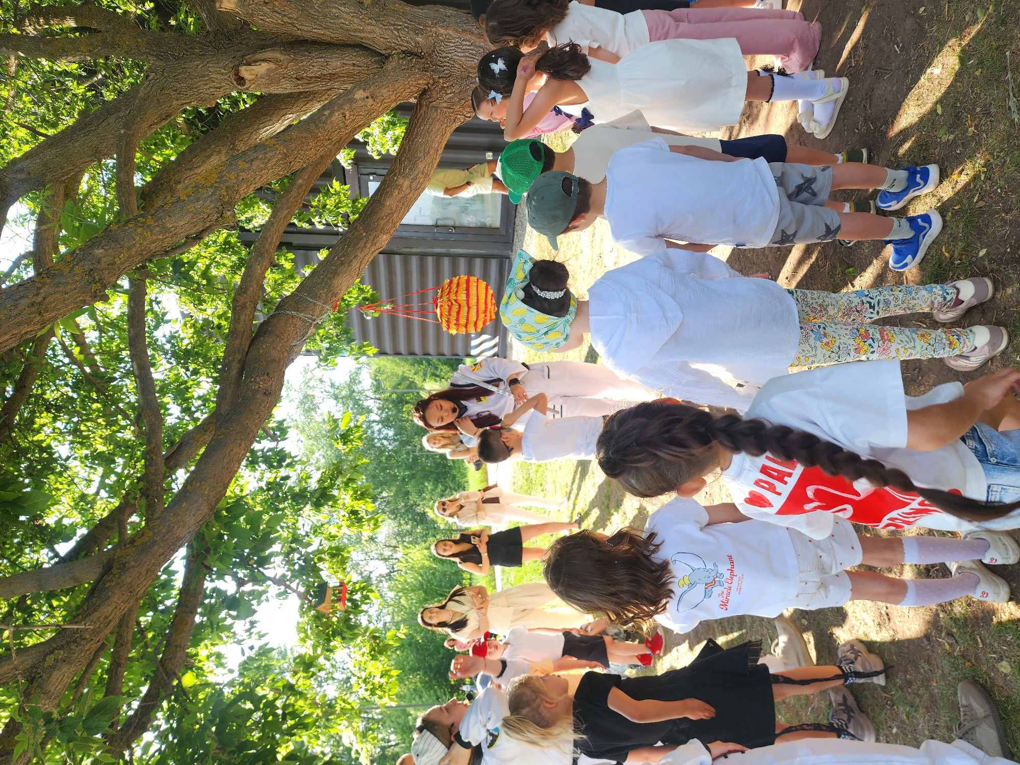 A group of children stand in a line, their arms outstretched, in an outdoor setting with a large tree overhead in Kazan, Russia. Most of the children are wearing white tops, and the rest of their clothing varies in color and pattern. A lantern hangs from the tree branches. The setting appears to be a park or a similar green space.