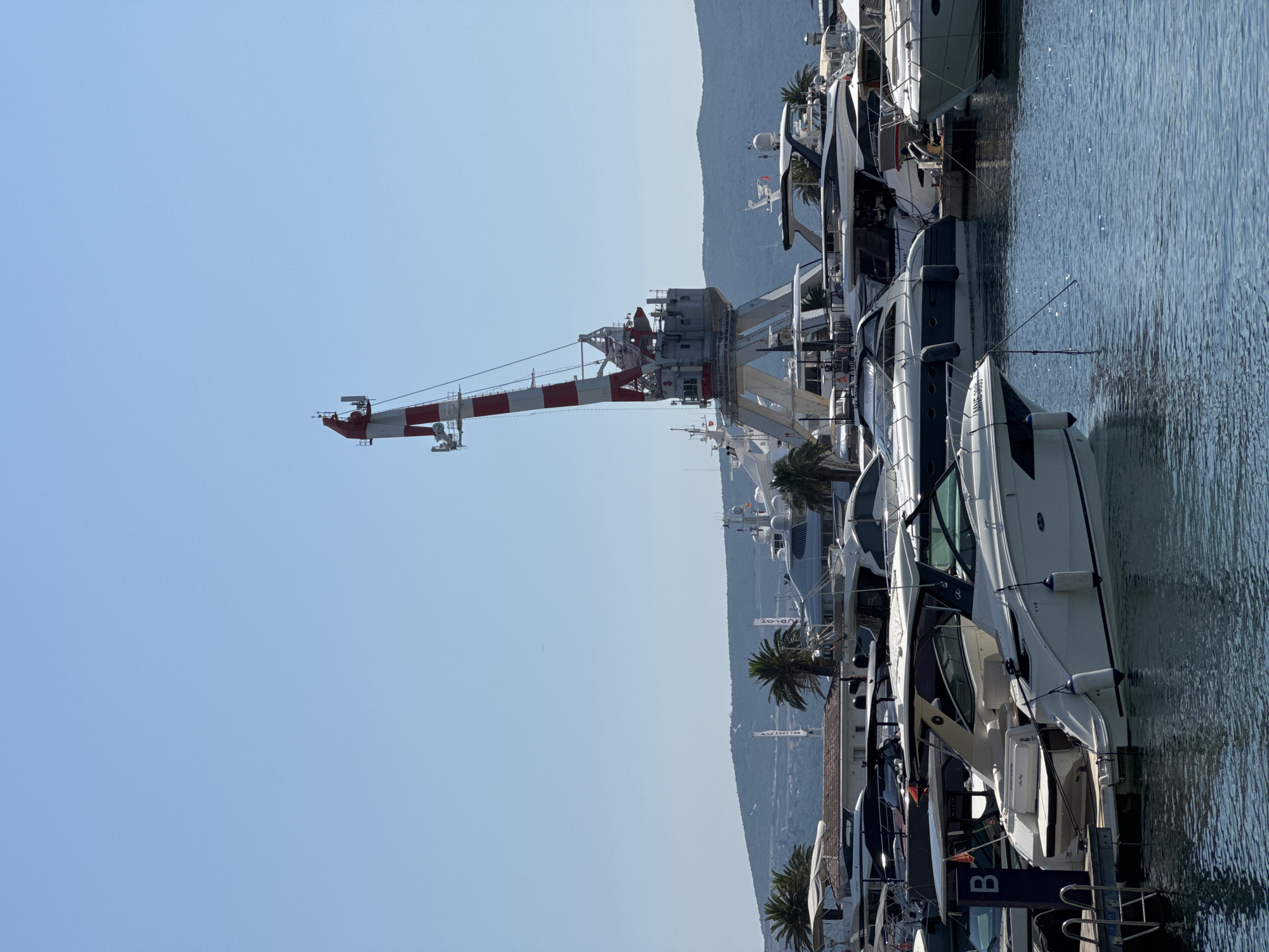 This is a photograph of a marina in Tivat, Montenegro. In the foreground, several white yachts are docked. One of the yachts has a sign that reads "B." The water in the marina is calm and reflects the bright blue sky. In the background, a large red and white crane stands tall over the marina. Palm trees and buildings are visible behind the yachts. A mountainous landscape can be seen in the distance. The overall scene depicts a sunny day at a modern marina.