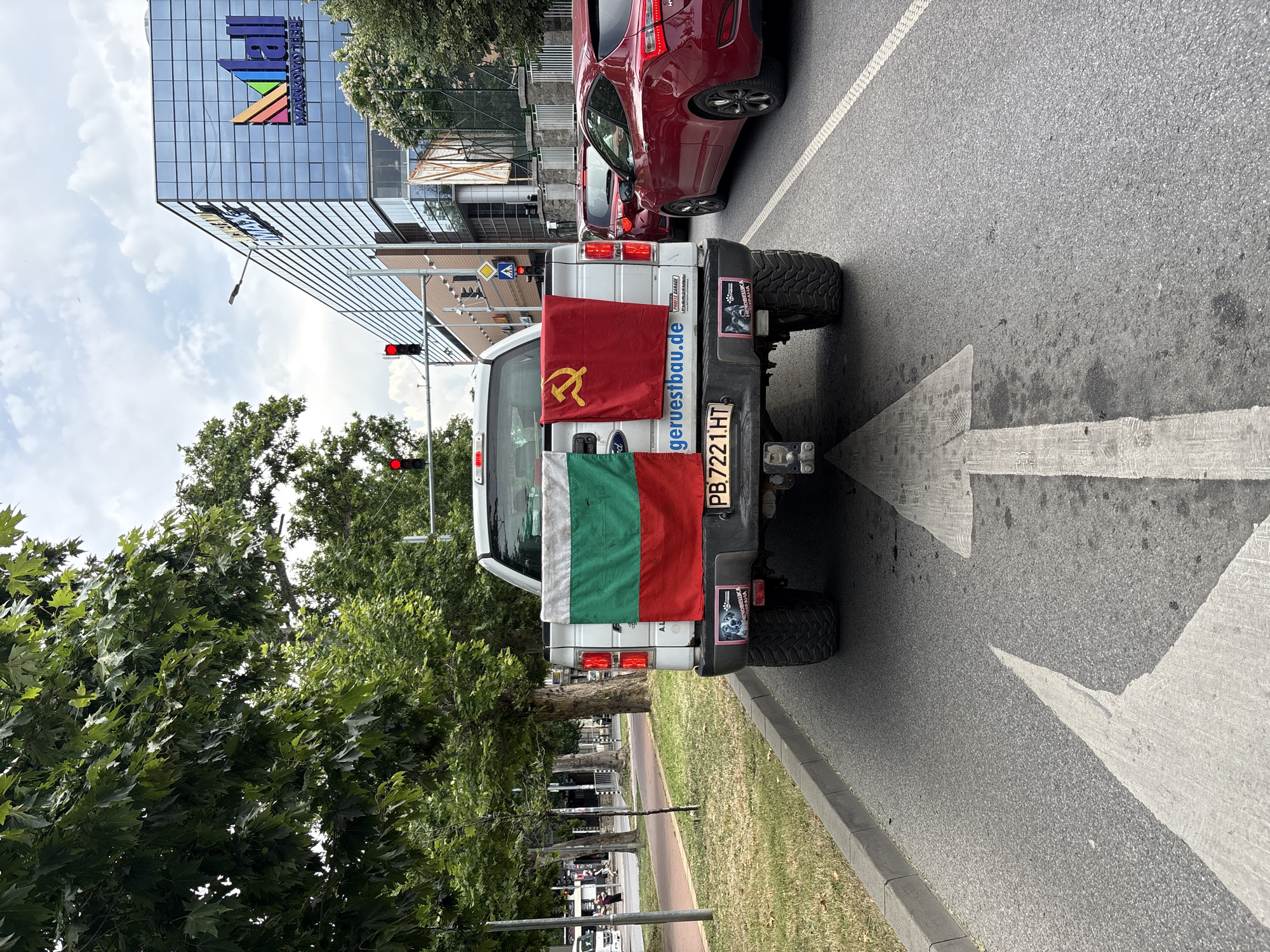 The image captures a street scene in Plovdiv, Bulgaria. In the foreground, the rear of a white pickup truck is prominent, adorned with two flags. The flag on the left is the national flag of Bulgaria, featuring horizontal stripes of white, green, and red. To its right, a red flag with a yellow hammer and sickle symbol, representing the Soviet Union, is displayed. The truck's license plate is visible as PB 7221 HT. Attached to the bumper are two stickers, one depicting a dog and text, and another with a stylized wolf-like creature. The truck is stopped on a paved road, indicated by a large white arrow painted on the asphalt, pointing forward.
Beyond the truck, the road continues, with other vehicles visible in traffic. A red car is in the lane to the right of the truck, and another red vehicle is further ahead. On the left side of the road, a sidewalk and grassy area are present, bordered by trees. In the background, a modern building with a reflective glass facade and a prominent "Mall" logo is situated. Traffic lights are visible on poles, with at least one showing a red signal. The sky is overcast with clouds. The overall scene depicts a street in an urban environment, likely during daylight hours, with the flags on the truck suggesting a political or nationalistic display.