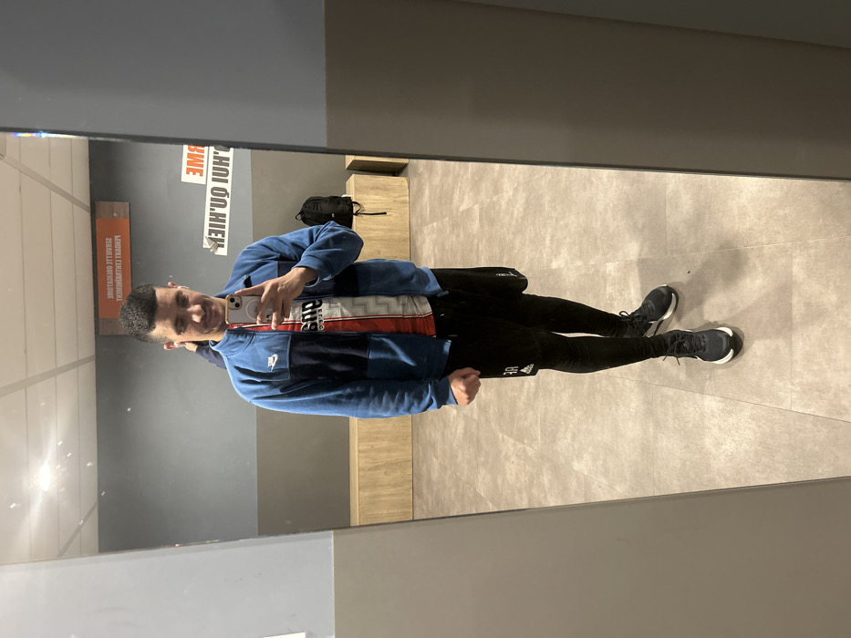A single male subject is depicted in a full-body mirror selfie taken within what appears to be a changing room or common area of a sports facility in Hœnheim, France. The subject has short, dark hair and a light complexion, exhibiting a slight smile. He is dressed in a blue and dark blue zippered athletic jacket with a white Nike logo on the left chest, worn over a white, red, and black diagonally striped athletic shirt or jersey featuring "PSG" branding. Black athletic shorts are worn over black compression leggings, and he wears dark athletic shoes with white soles. His right hand holds a light-colored smartphone, positioned to capture the image, while his left hand is clenched into a fist.
The environment, visible in the mirror's reflection, consists of a grey wall with a light wood-paneled bench extending along its base and a light grey tiled floor. A black backpack rests on a section of the bench to the subject's right. An orange rectangular sign is affixed to the wall, displaying inverted white text, partially legible as "SERVIETTE OBLIGATOIRE" (Towel Required), indicating a facility rule. Overhead lighting is reflected in the mirror.
