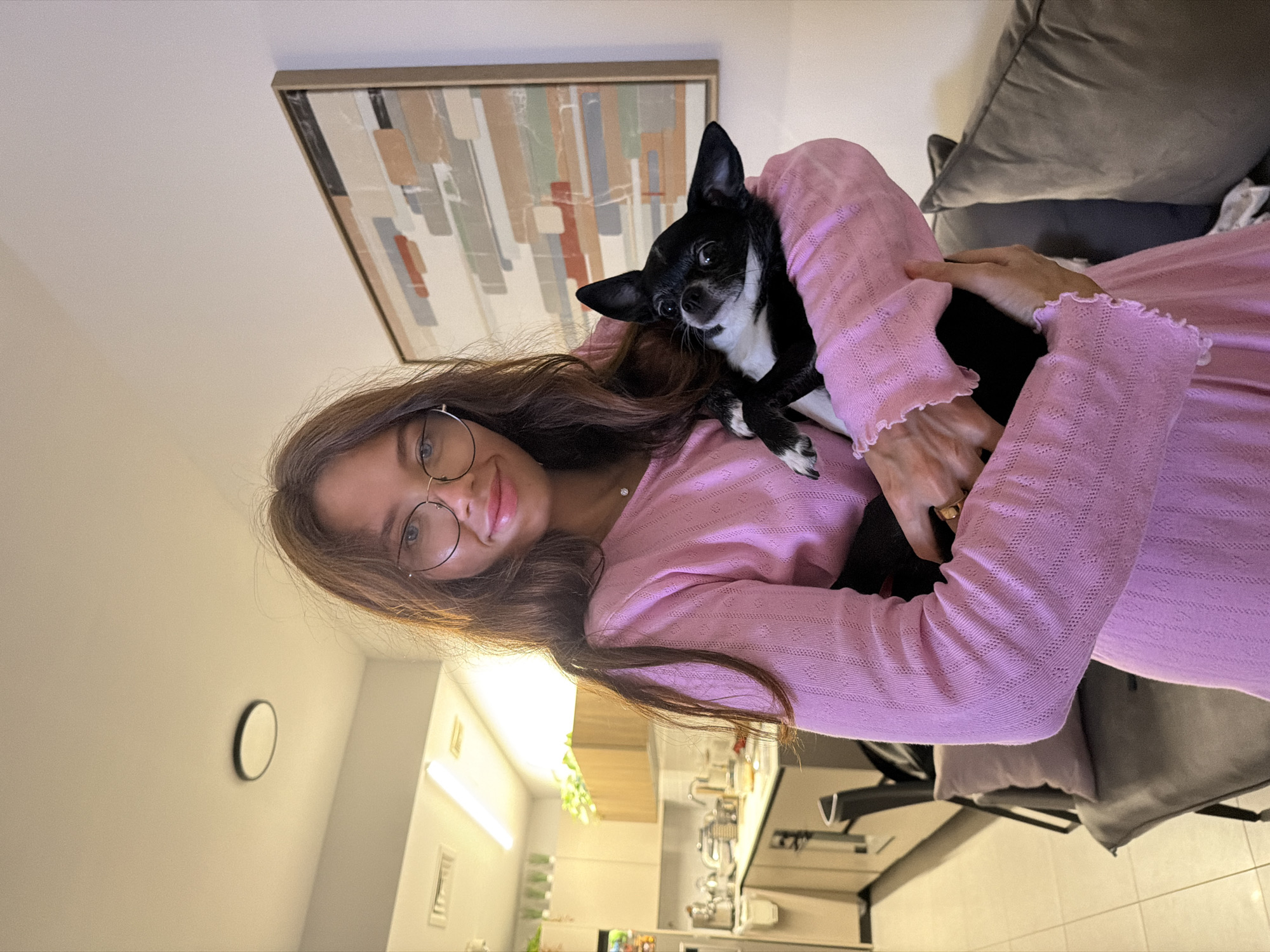 A young woman with long brown hair and glasses holds a black and white chihuahua dog. She is wearing a pink, long-sleeved, patterned sweater. Behind them is an abstract painting in a neutral frame. The background shows part of a kitchen with white cabinets and appliances, suggesting an apartment interior. A round clock is visible on the wall in the background. The location cannot be verified as Tel-Aviv, Israel, from the image alone.